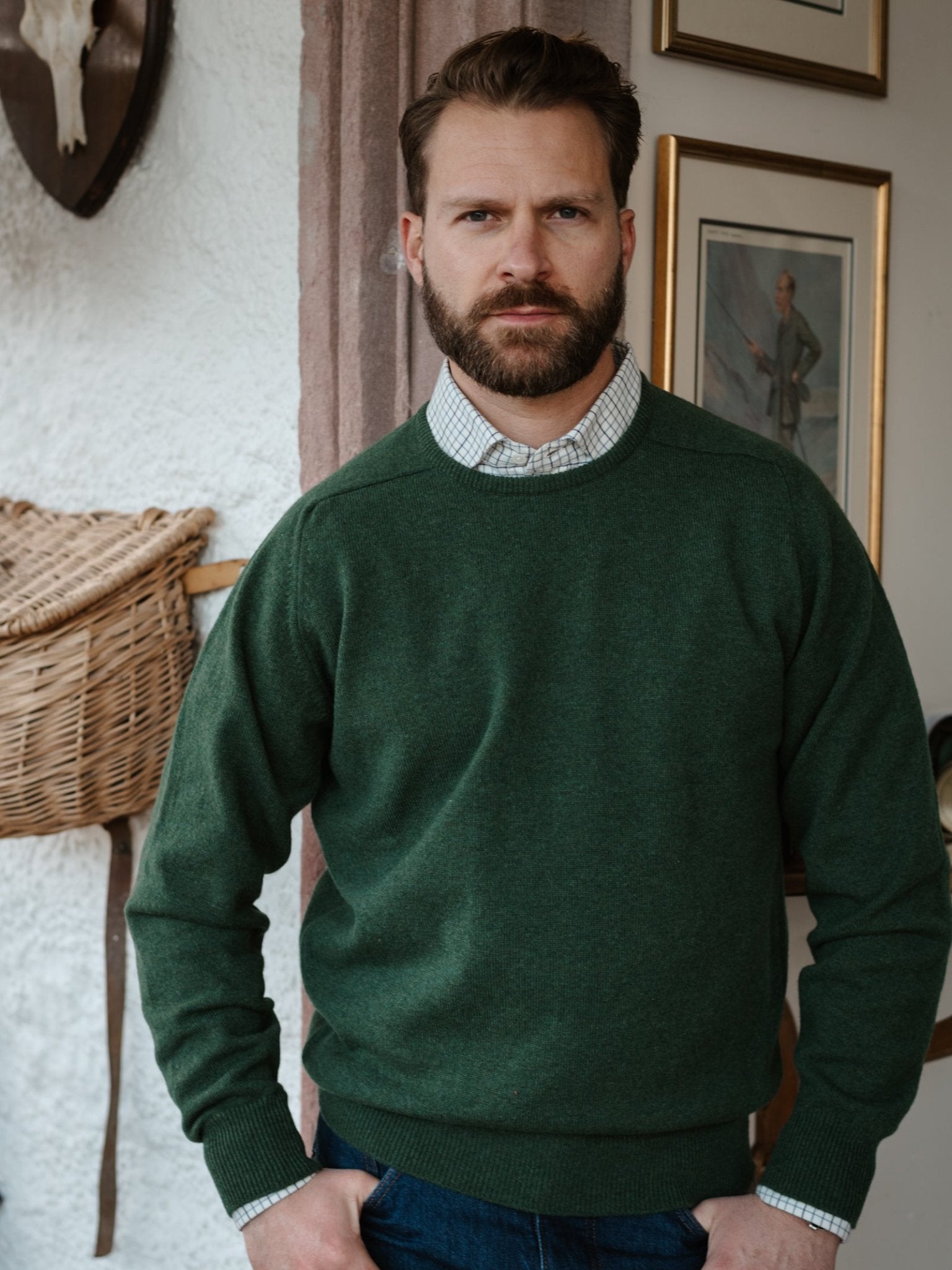 A man with short brown hair and a beard wears the Campbell's of Beauly Lambswool Crew Neck Jumper over a checked shirt, standing indoors with framed pictures and a wicker basket on the wall behind him.