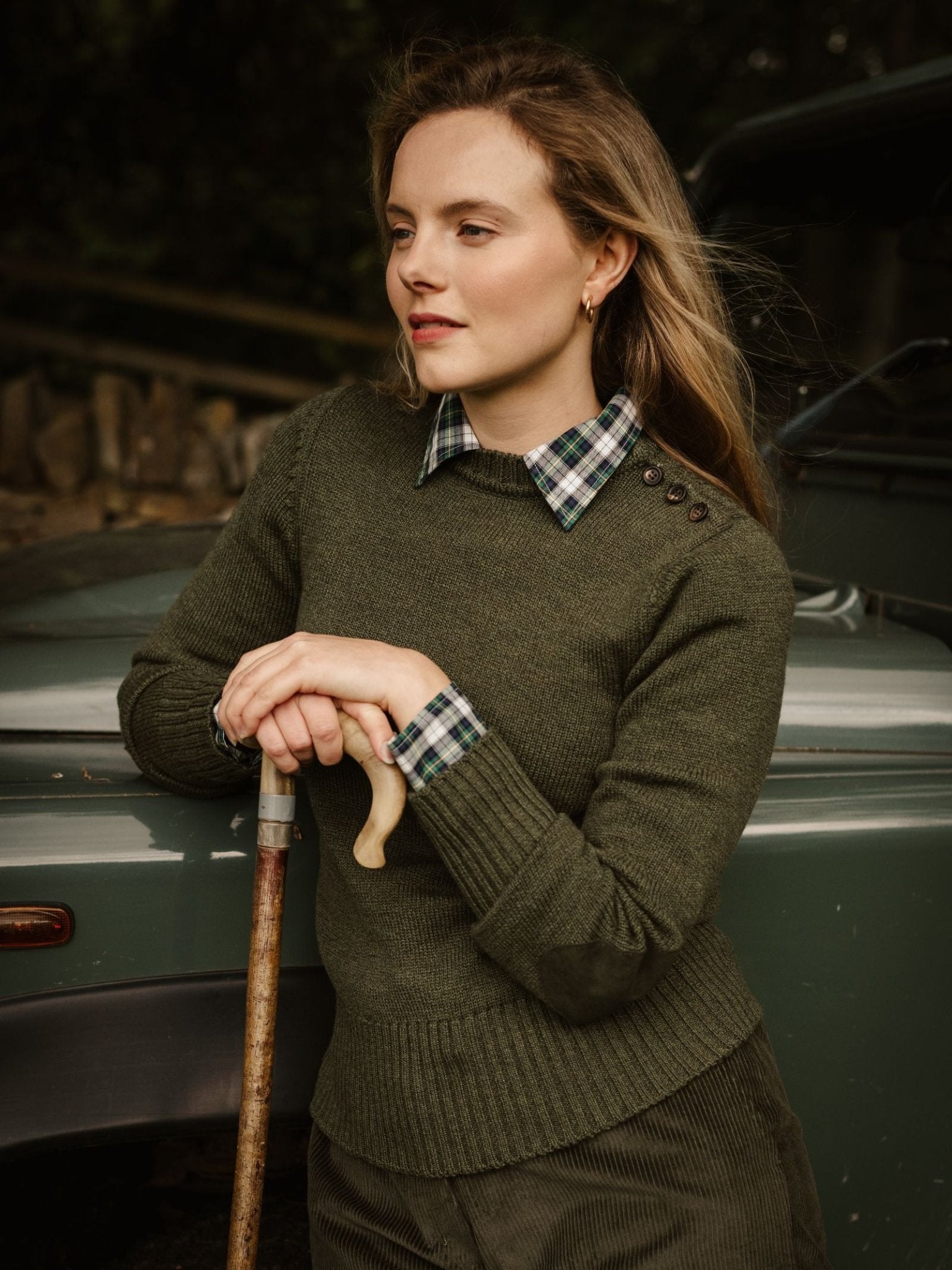 A woman with light brown hair and a calm expression wears a Campbell’s of Beauly Lambswool Crew Neck Shooter Jumper in olive green, layered over a plaid collared shirt, leaning on a cane before a green car outdoors.