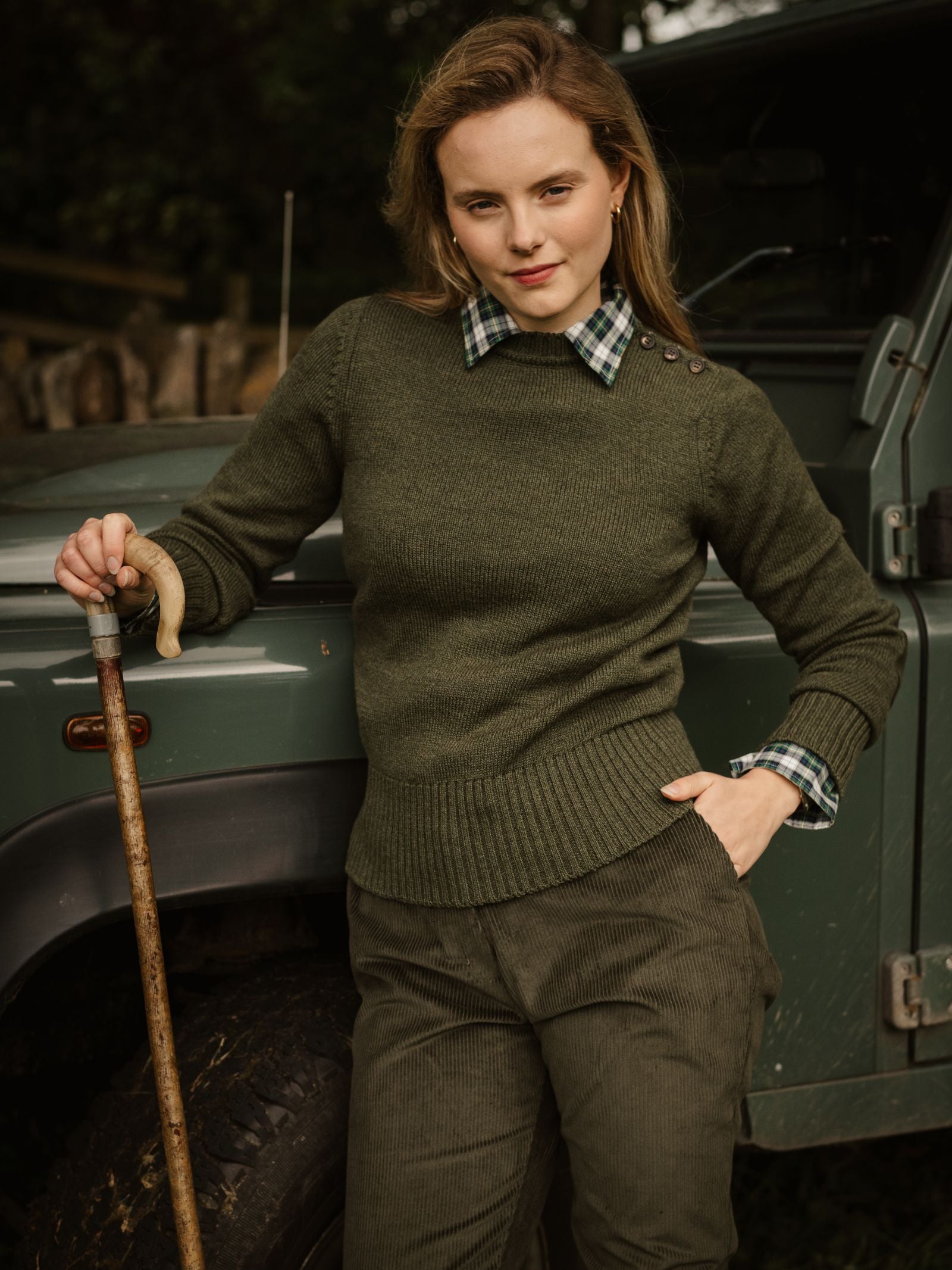 A woman with light hair and a confident expression stands outdoors in a Campbell's of Beauly Lambswool Crew Neck Shooter Jumper, layered over a plaid shirt with green corduroy pants, leaning on a wooden stick beside a green off-road vehicle.