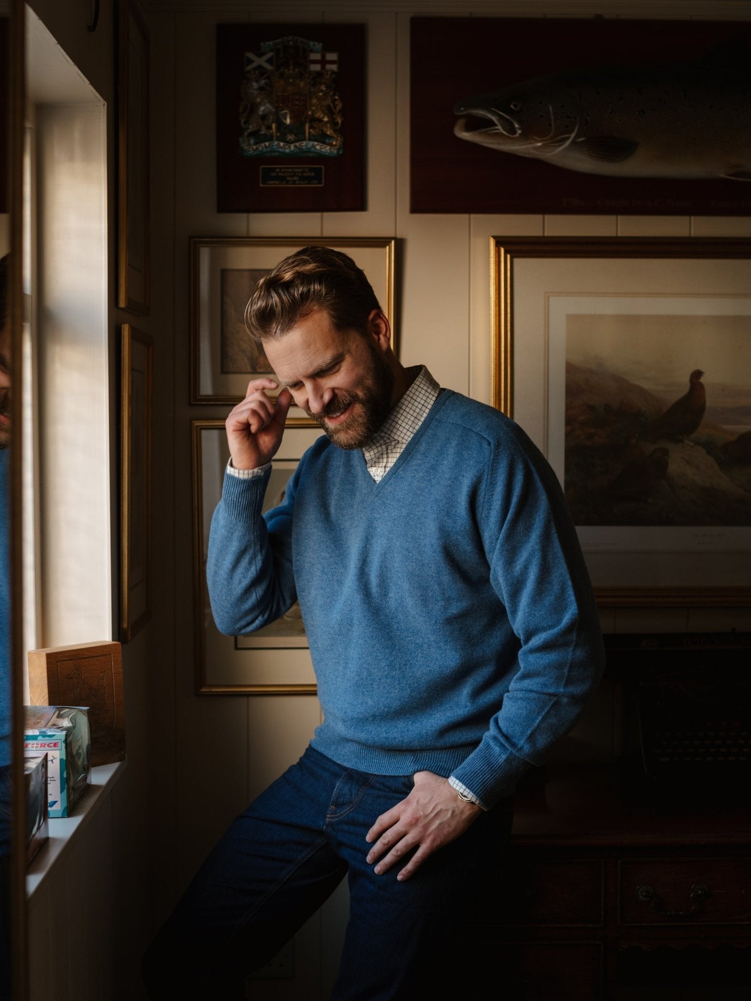 A man in a Campbell's of Beauly Lambswool V-Neck Jumper and jeans stands by a window, smiling and touching his temple. The cozy room features framed pictures, a large fish model, and warm lighting.