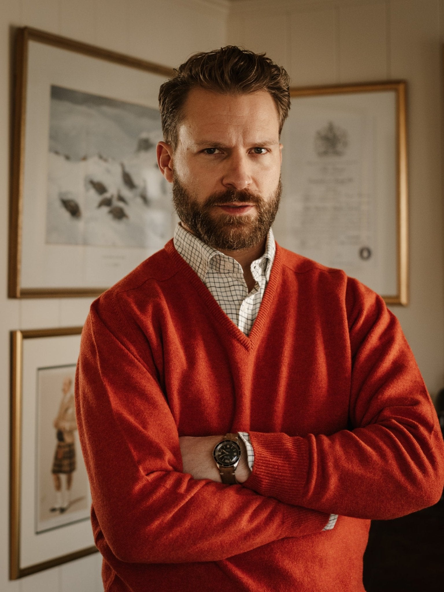 A bearded man with a mustache wearing the Campbell's of Beauly Lambswool V-Neck Jumper over a checkered shirt stands with his arms crossed in front of framed artwork on the wall.