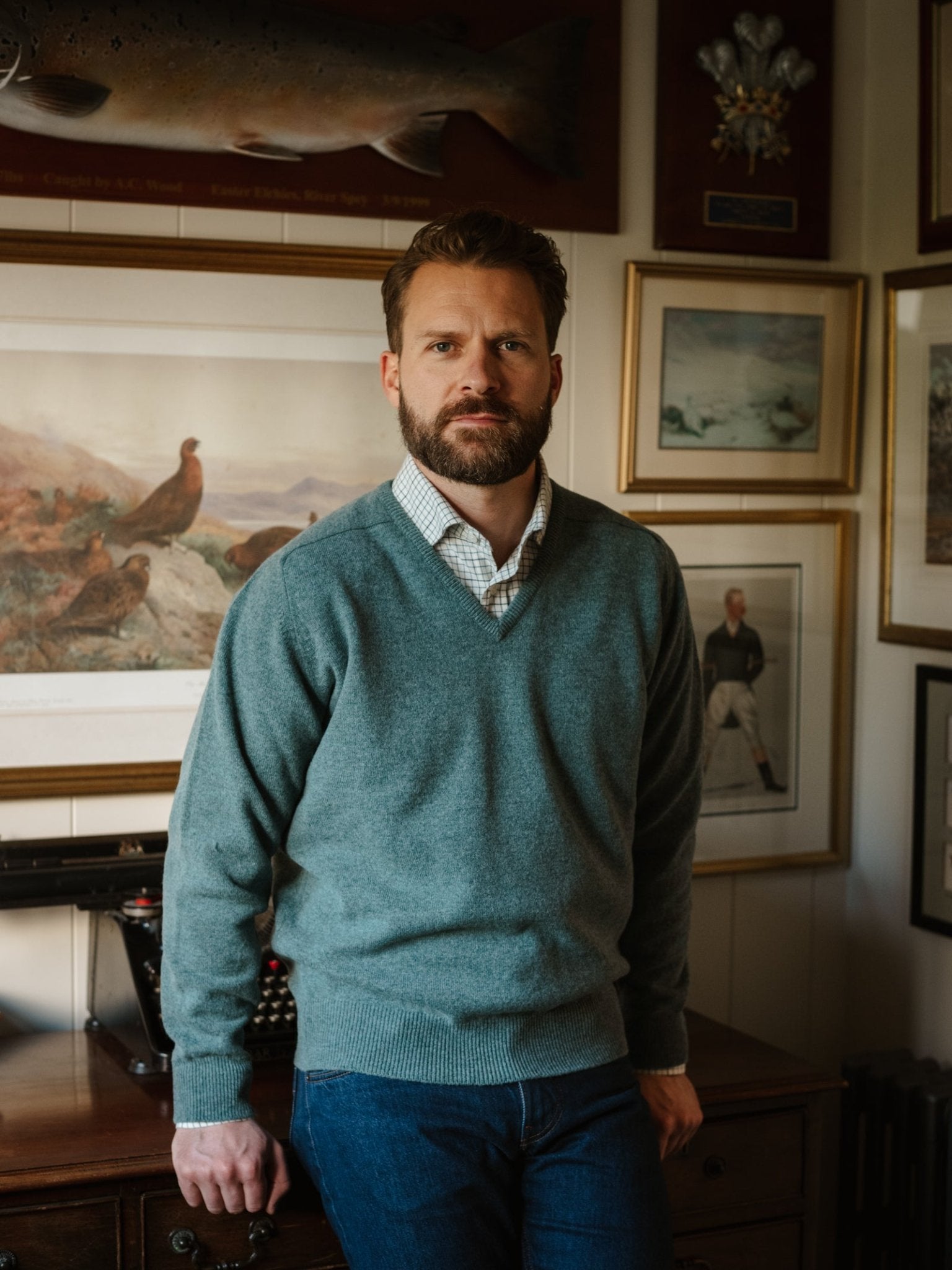 A bearded man in a classic-fit Campbell's of Beauly Lambswool V-Neck Jumper and jeans stands indoors, leaning on a wooden desk. Behind him, the wall features framed artwork such as wildlife prints and a mounted fish.