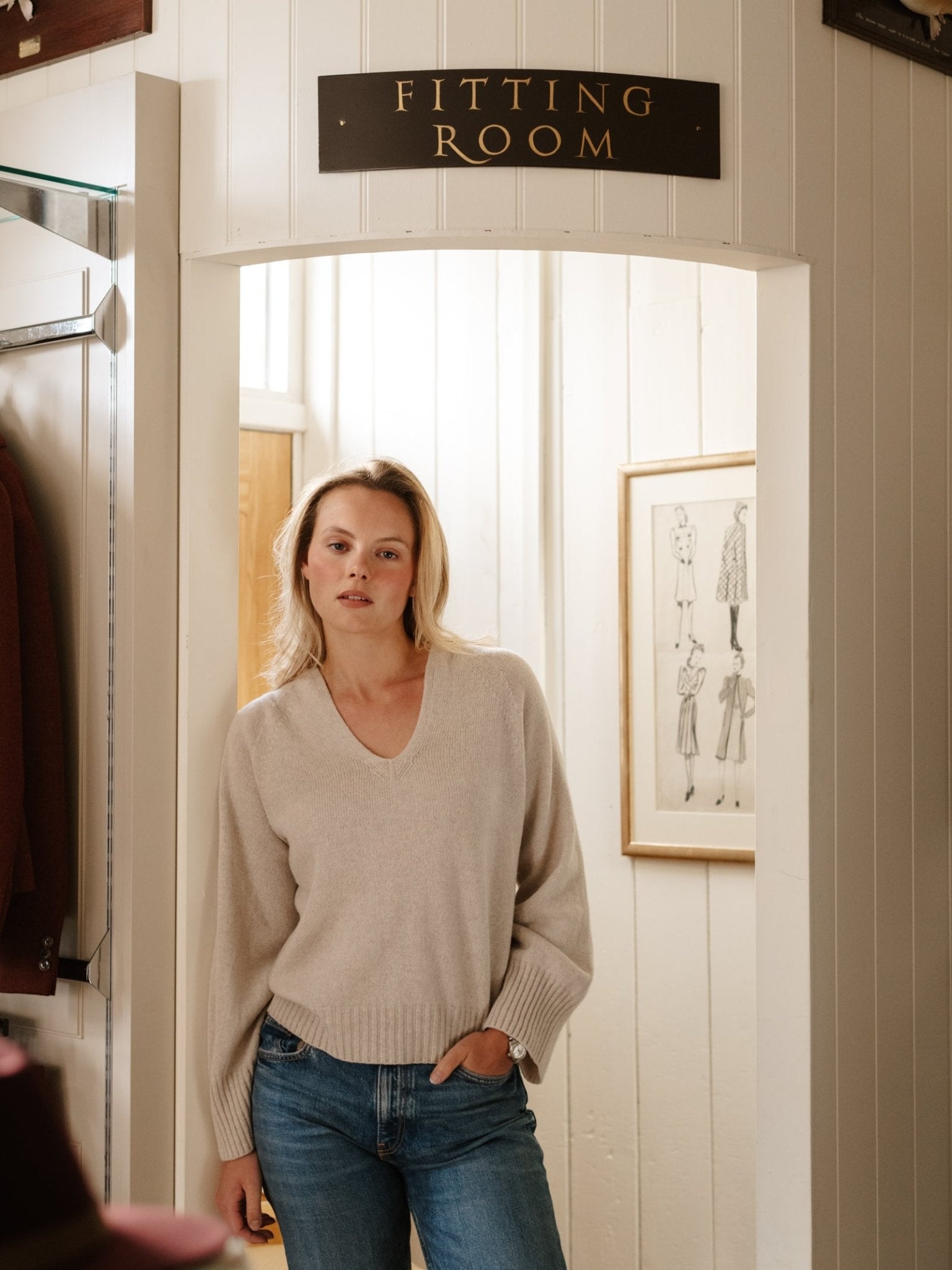 A woman in a beige Campbell's of Beauly Geelong Lambswool V Jumper and blue jeans stands in a doorway beneath a "Fitting Room" sign, with framed fashion sketches visible on the wall behind her.