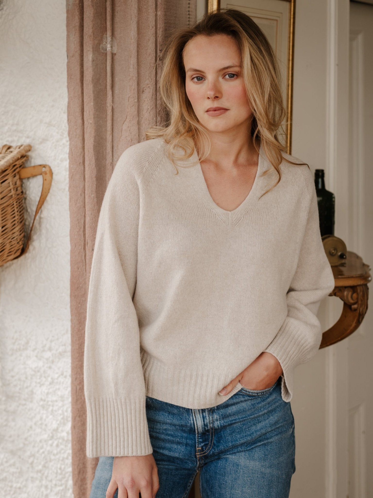 A woman with wavy blonde hair wears a light beige Geelong Lambswool V Jumper by Campbell's of Beauly and blue jeans, standing indoors near a basket on the wall and a door, with a neutral expression.
