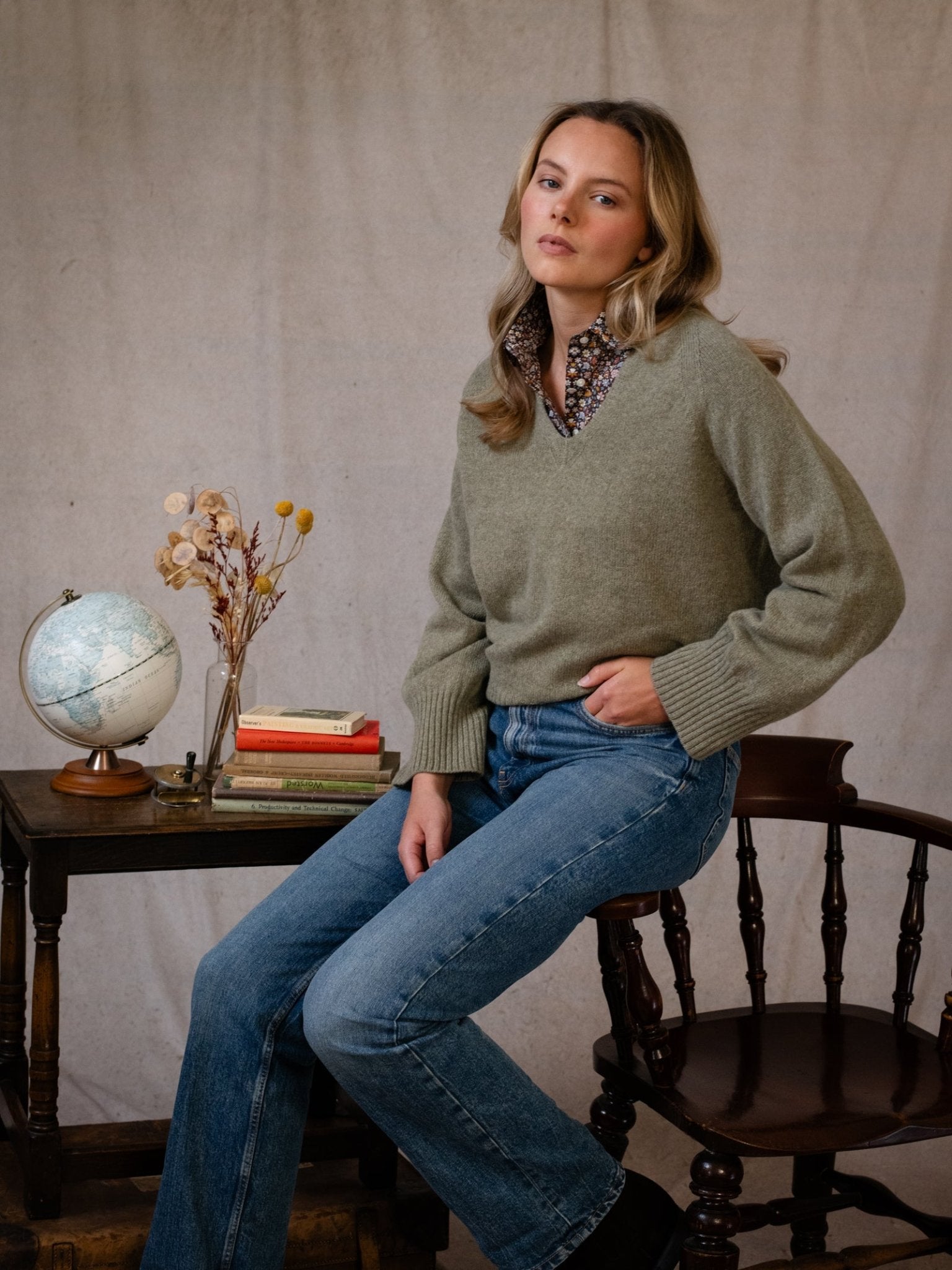 A woman in a Campbell's of Beauly Geelong Lambswool V Jumper (green) and blue jeans sits on the arm of a dark wooden chair beside a small table with a globe, books, and dried flowers, set against a plain beige backdrop.