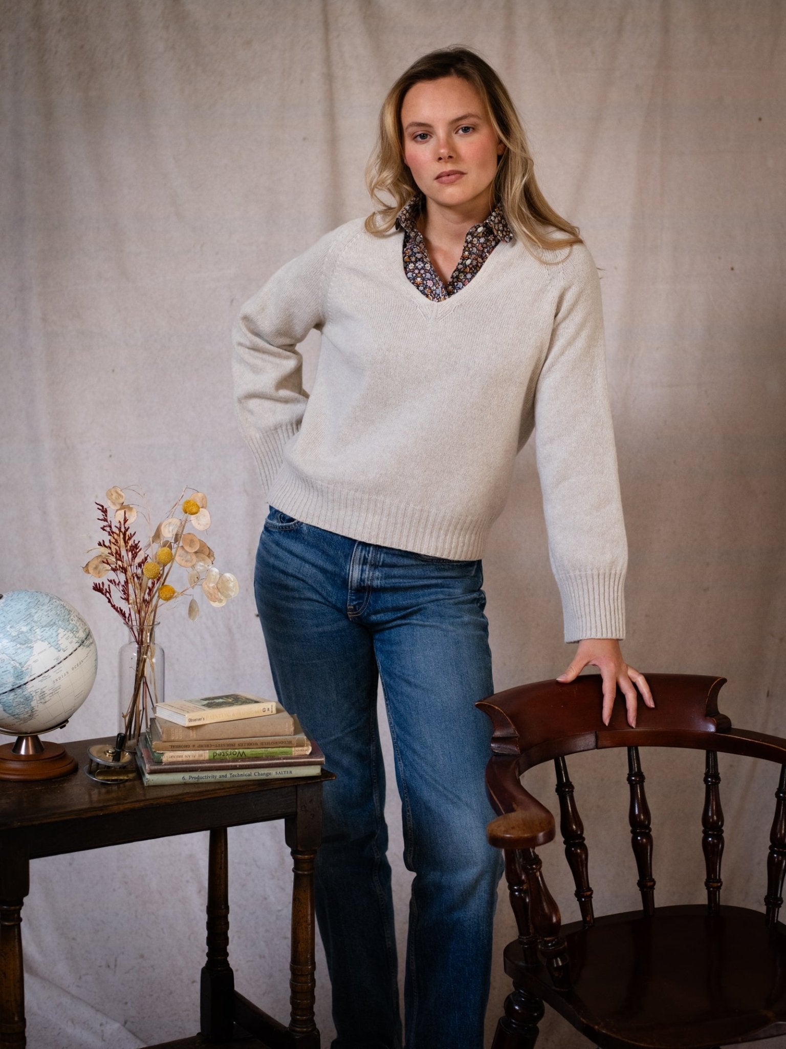 A woman in a Campbell's of Beauly Geelong Lambswool V Jumper and jeans stands beside a wooden chair and table with books, a globe, and dried flowers against a neutral fabric backdrop.
