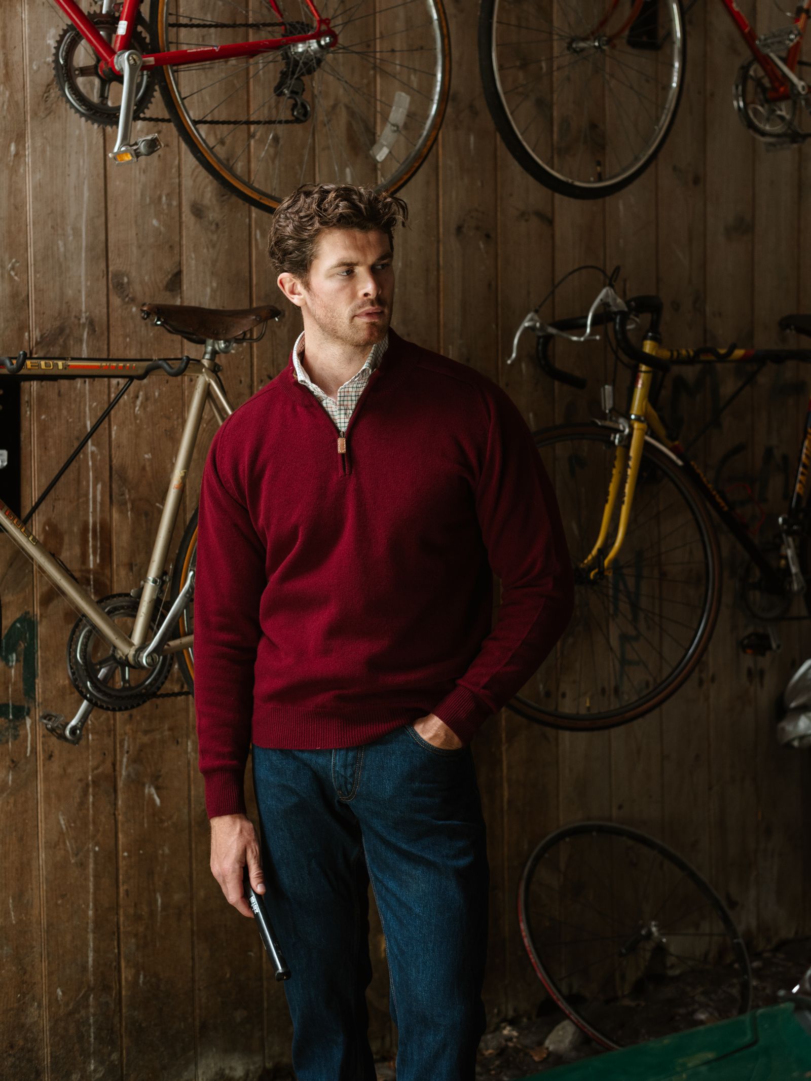 A man wearing a Campbell's of Beauly Lambswool Quarter Zip Jumper and blue jeans stands in a wooden room with wall-mounted bicycles behind him, one hand in his pocket as he gazes thoughtfully to the side.