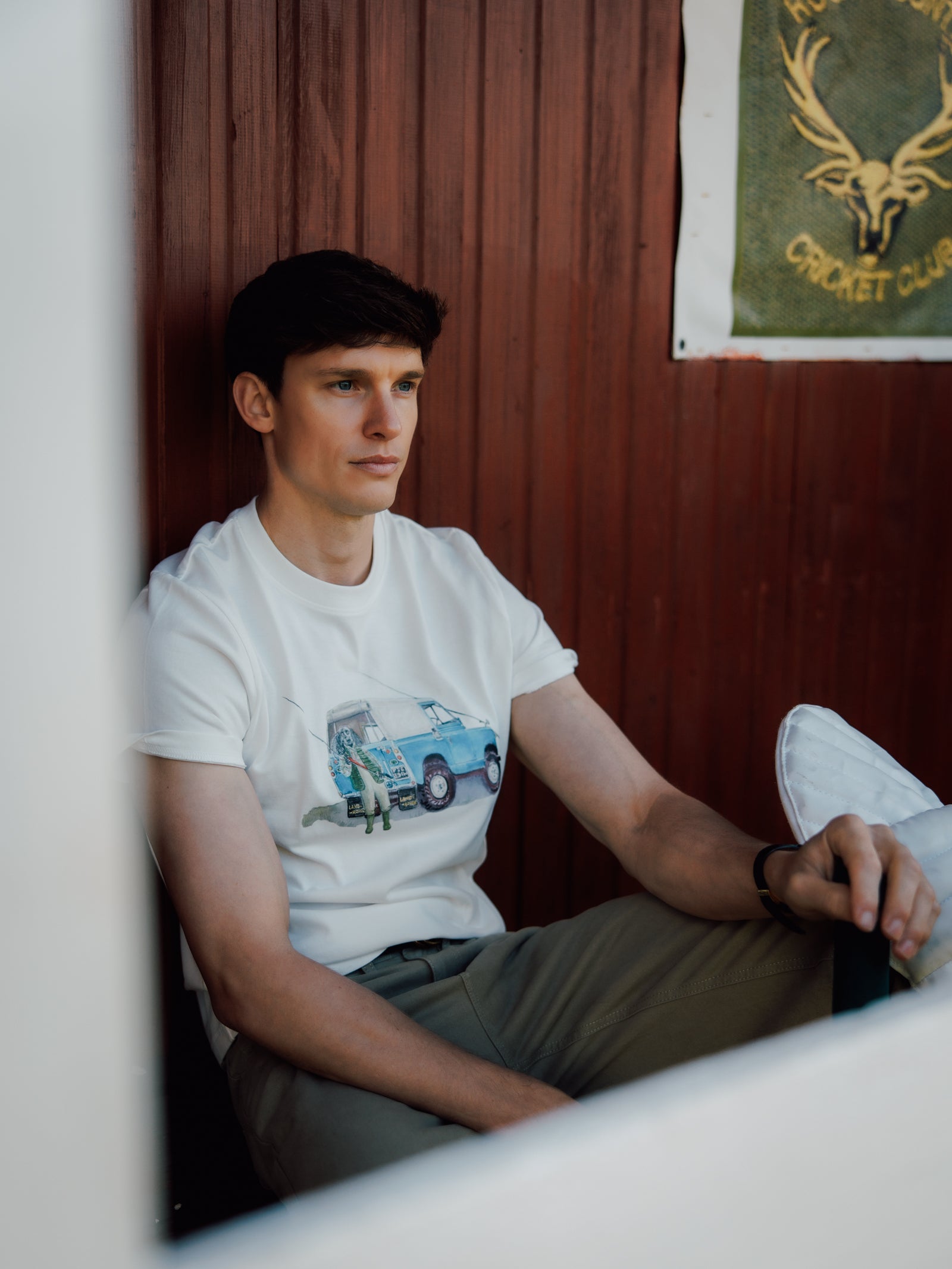 A man with short dark hair sits against a wooden wall, wearing khaki pants and a white Campbell's of Beauly Land Rover Cocker T-Shirt featuring a graphic. He gazes to the side, with part of a green sign bearing a deer logo visible in the background.