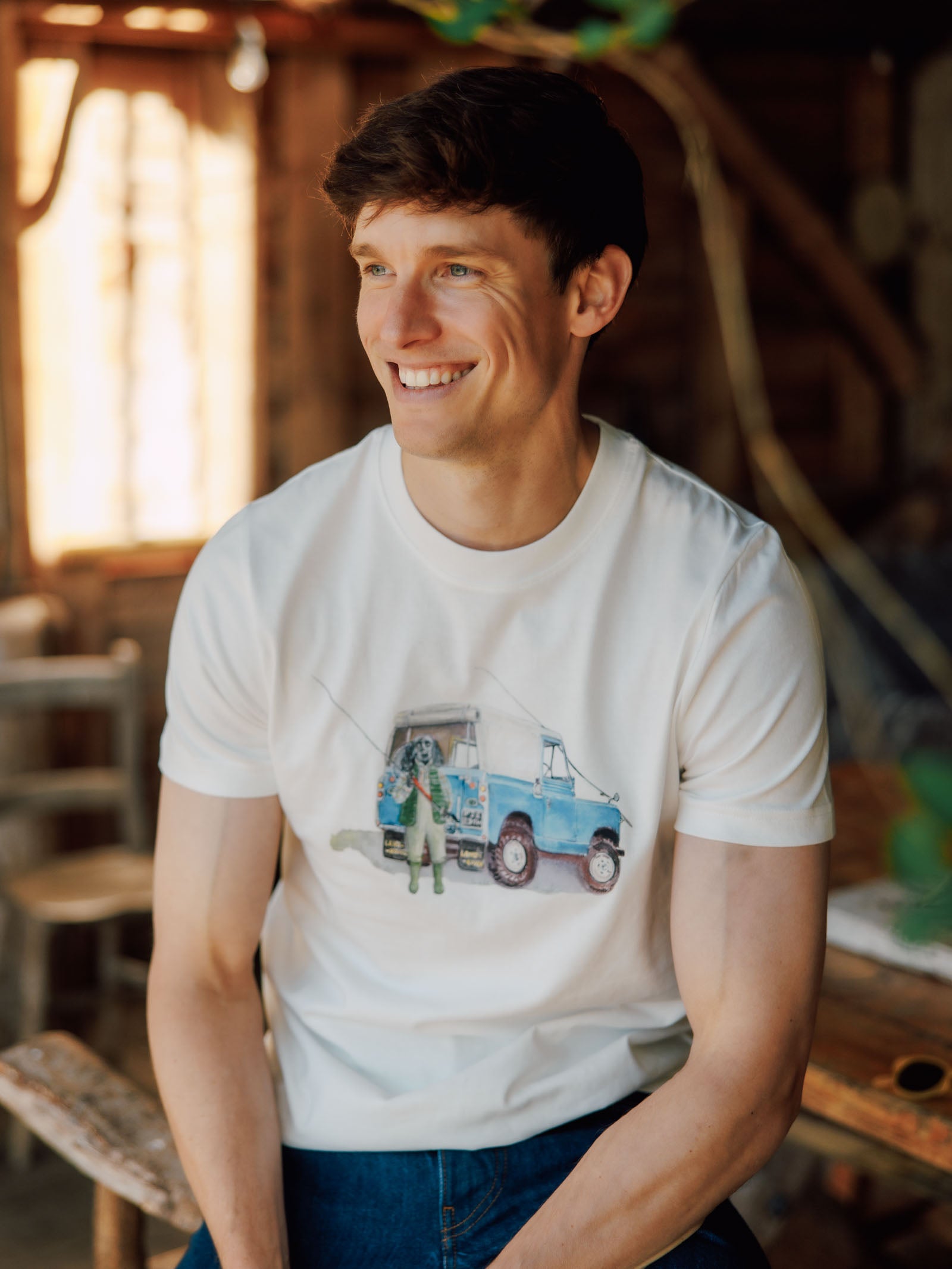 A smiling man with short brown hair wears the Campbell's of Beauly Land Rover Cocker T-Shirt, featuring a colorful blue van illustration, as he sits indoors on a wooden chair with sunlight streaming through the window behind him.
