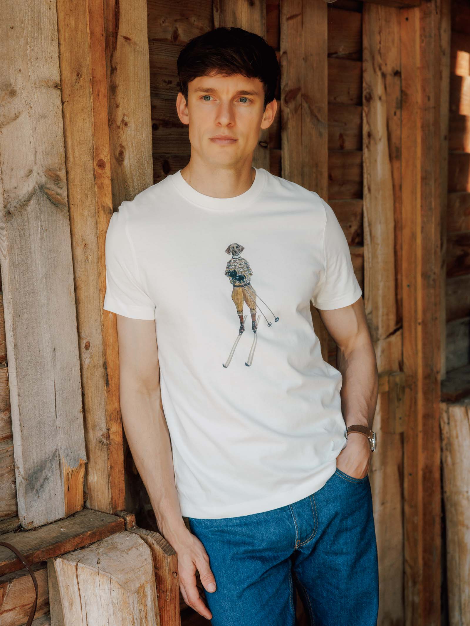 A man with brown hair wears the Campbell's of Beauly Le Ski Labrador T-Shirt—a cotton graphic tee—and blue jeans, standing casually against a wooden wall with one hand in his pocket.