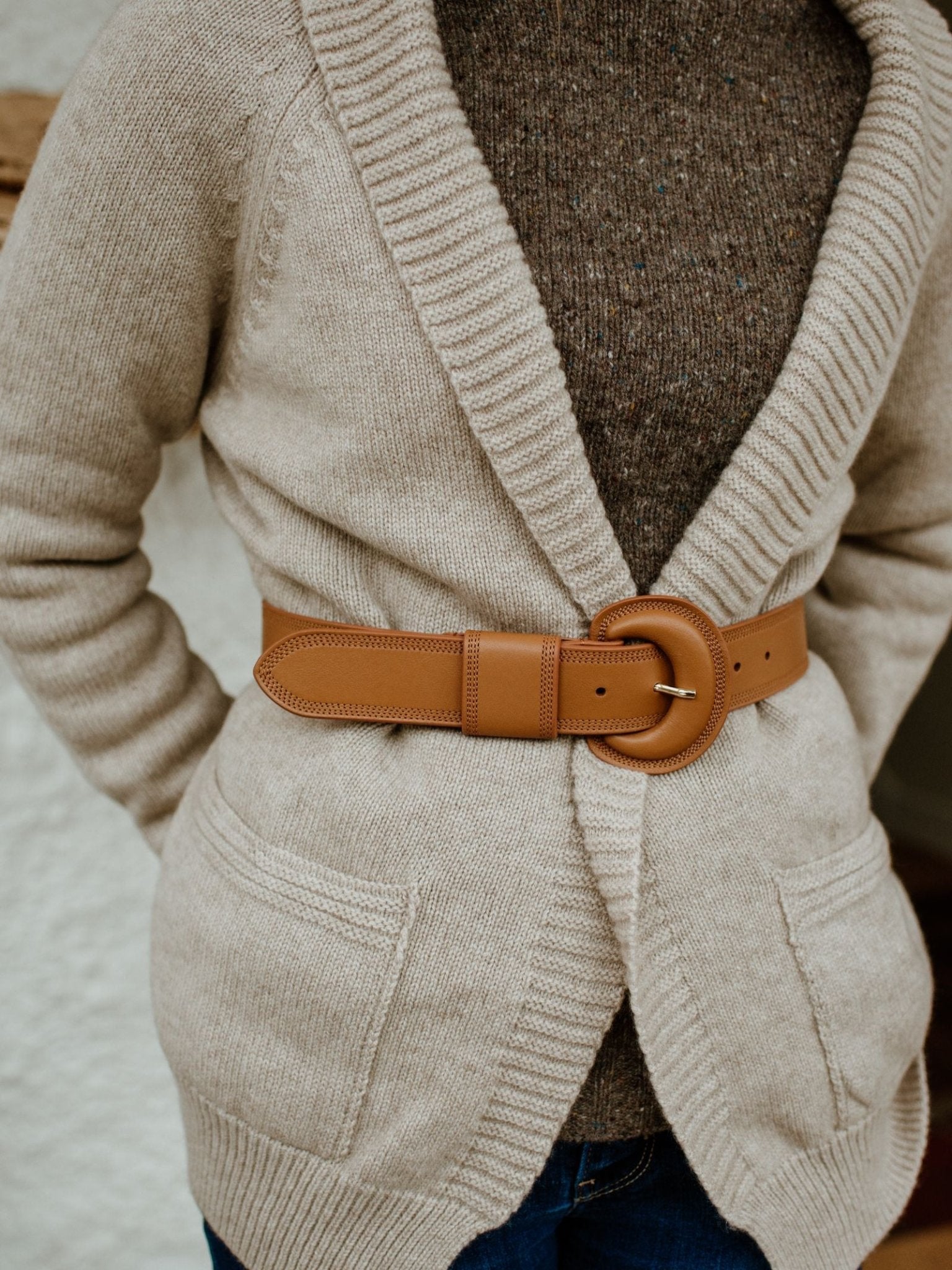 Wearing a beige knit cardigan with large pockets, cinched at the waist with the Caramel Leather Belt by Campbell's of Beauly featuring a brass buckle, layered over a dark brown sweater and blue jeans.