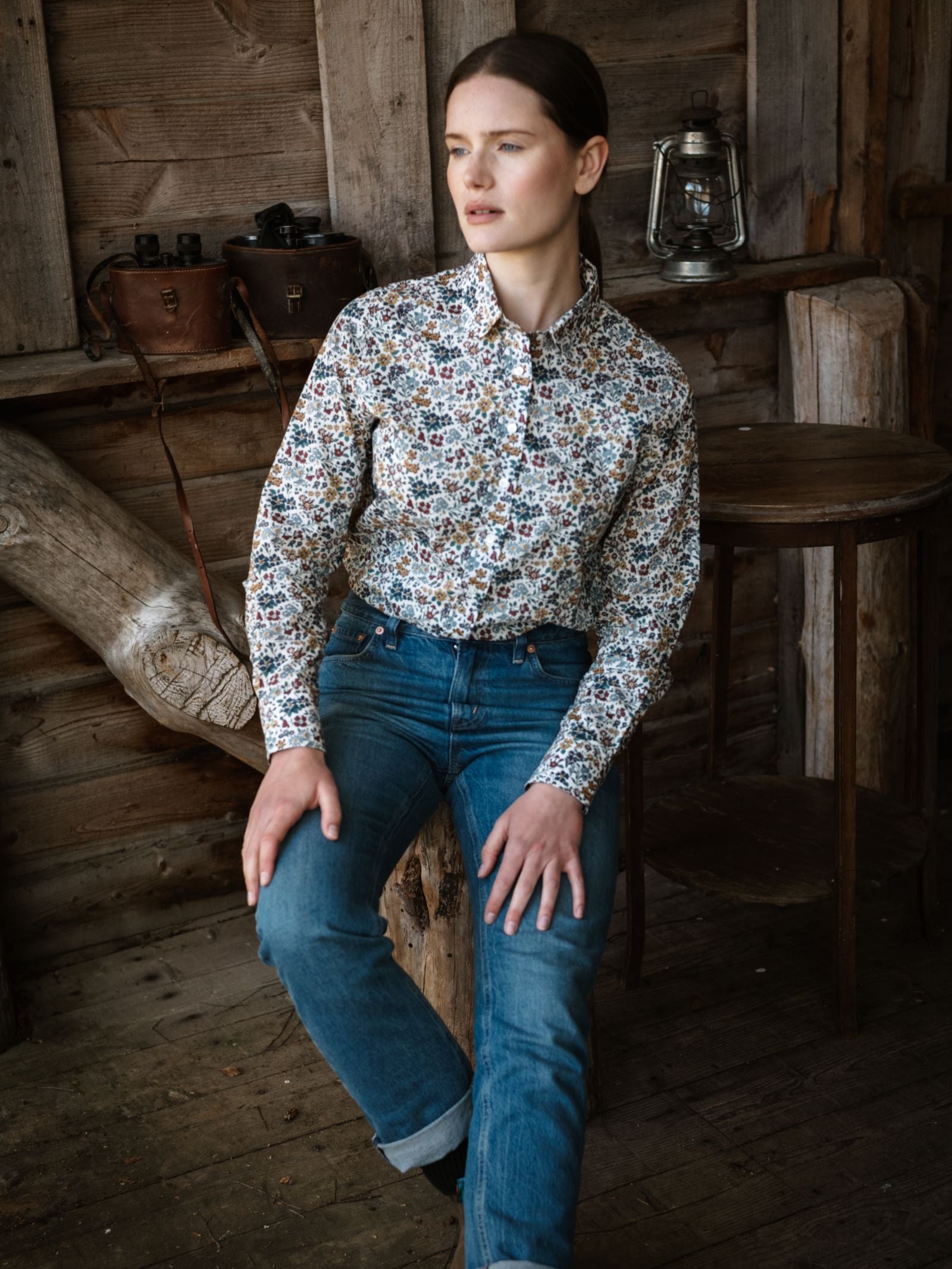 A woman wearing a Campbell's of Beauly Liberty Print Cotton Shirt and blue jeans sits on a wooden bench in a rustic room with wood-paneled walls, a round table, an old lantern, and leather saddlebags, gazing thoughtfully to the side.