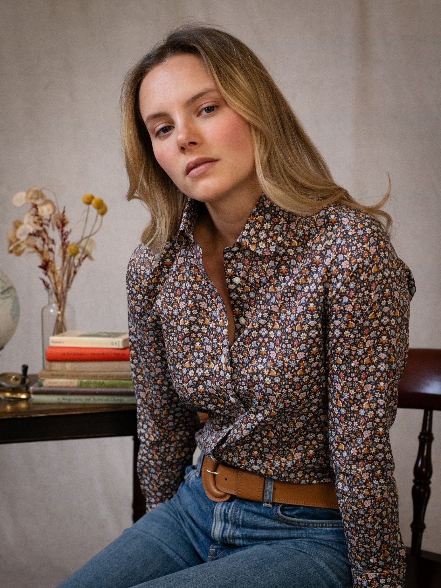 A woman with long blonde hair wears a Campbell's of Beauly Liberty Print Cotton Shirt and jeans, sitting on a chair beside a table with stacked books, dried flowers, and a globe, looking calmly at the camera.