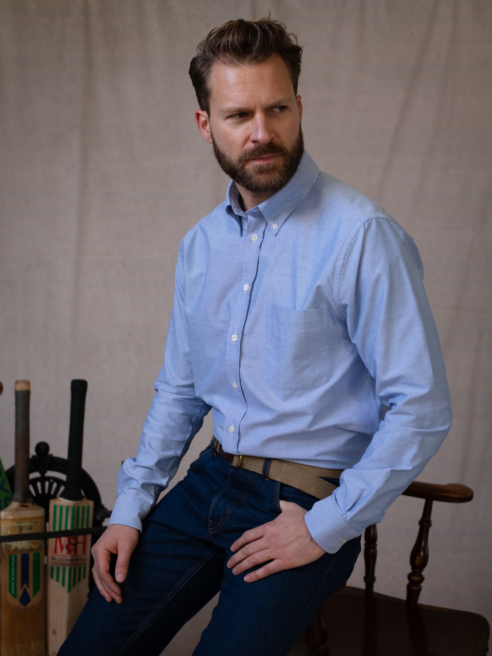 A bearded man sits on a wooden chair, wearing a light blue Campbell's of Beauly Oxford Button Down Shirt and dark jeans. He looks to the side with a serious expression, with cricket bats and equipment in the background.