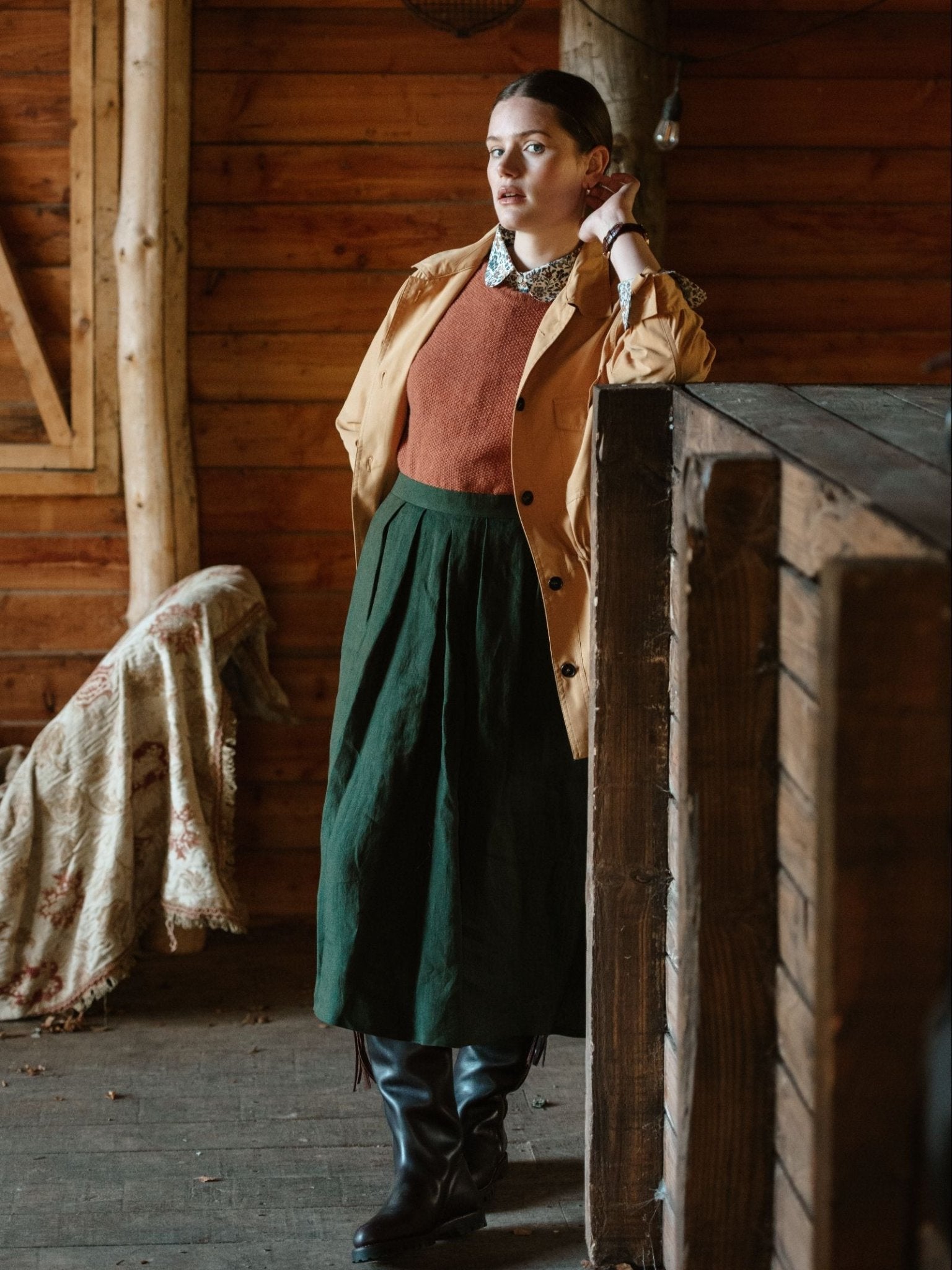 Wearing a Campbell's of Beauly Lightweight Aigas Field Jacket, rust sweater, green skirt, and black boots, a person stands indoors by a wooden wall and counter, touching their neck and gazing thoughtfully ahead.
