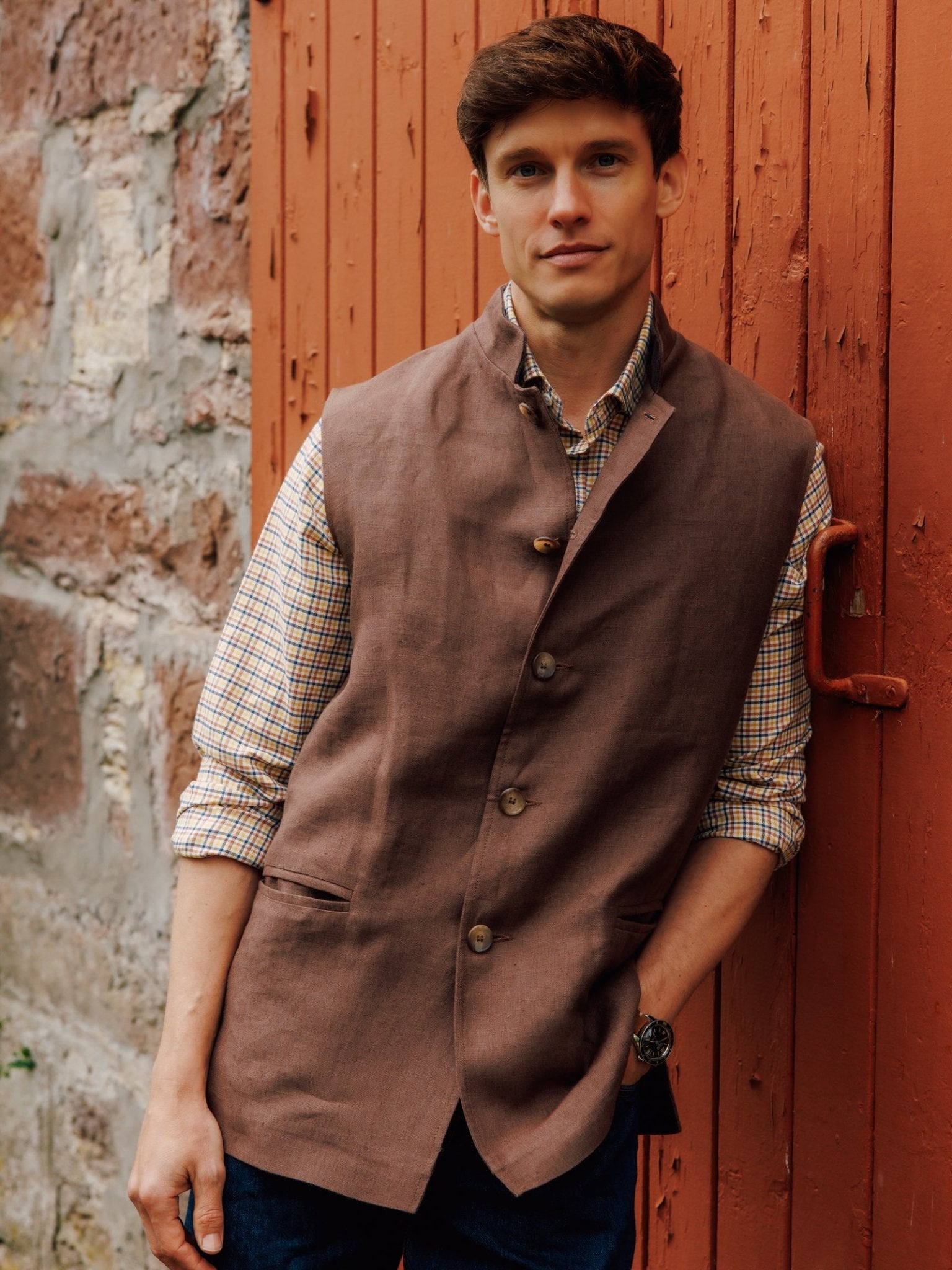 A man with short brown hair wears a Campbell's of Beauly Linen Nehru Gilet over a checked shirt and jeans, standing by a reddish wooden door and stone wall. One hand is in his pocket, his expression neutral.