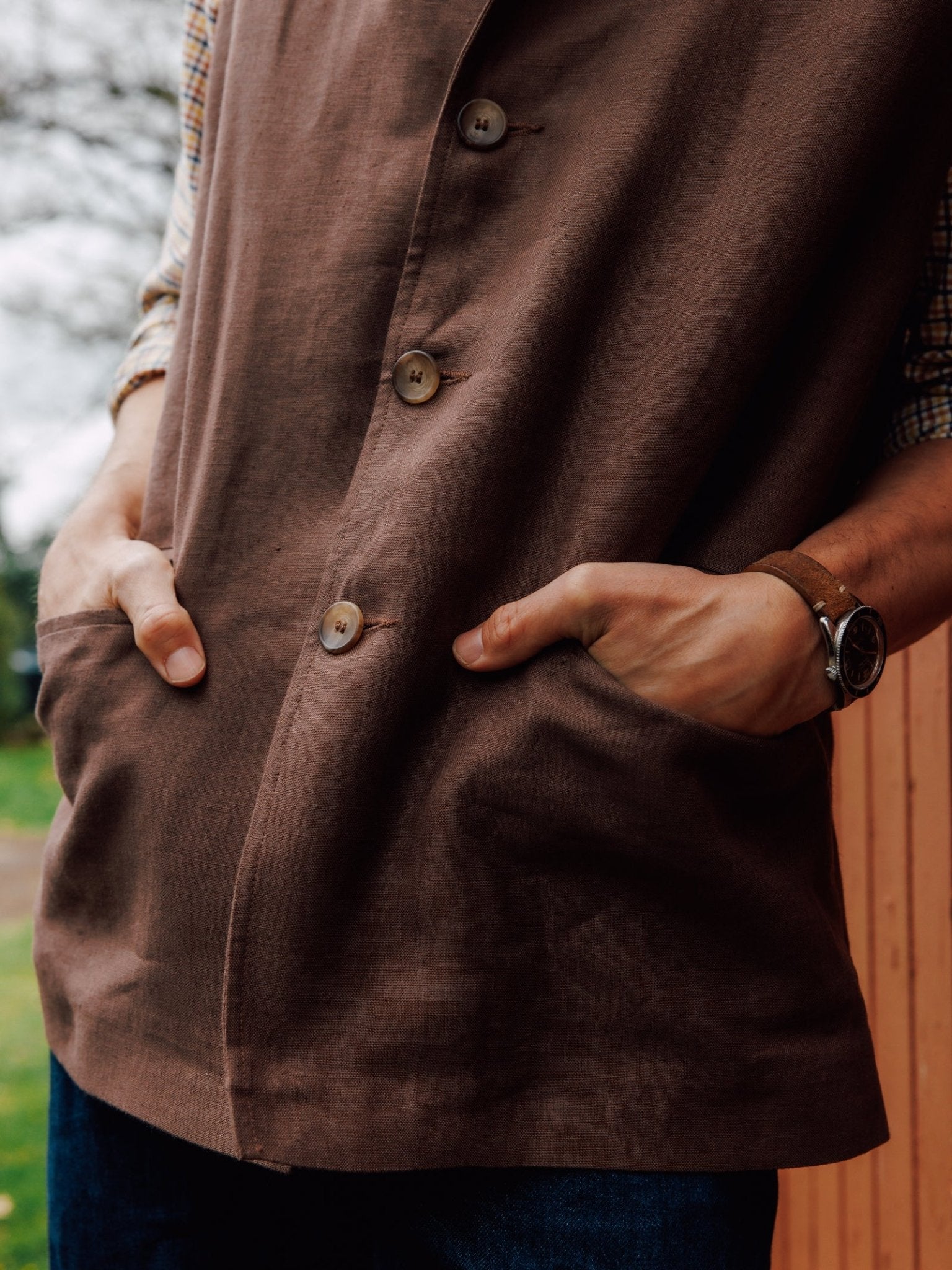 A person stands outdoors by a wooden wall, hands in pockets, wearing a Campbell's of Beauly Linen Nehru Gilet layered over a plaid shirt, with blue jeans and a brown wristwatch.