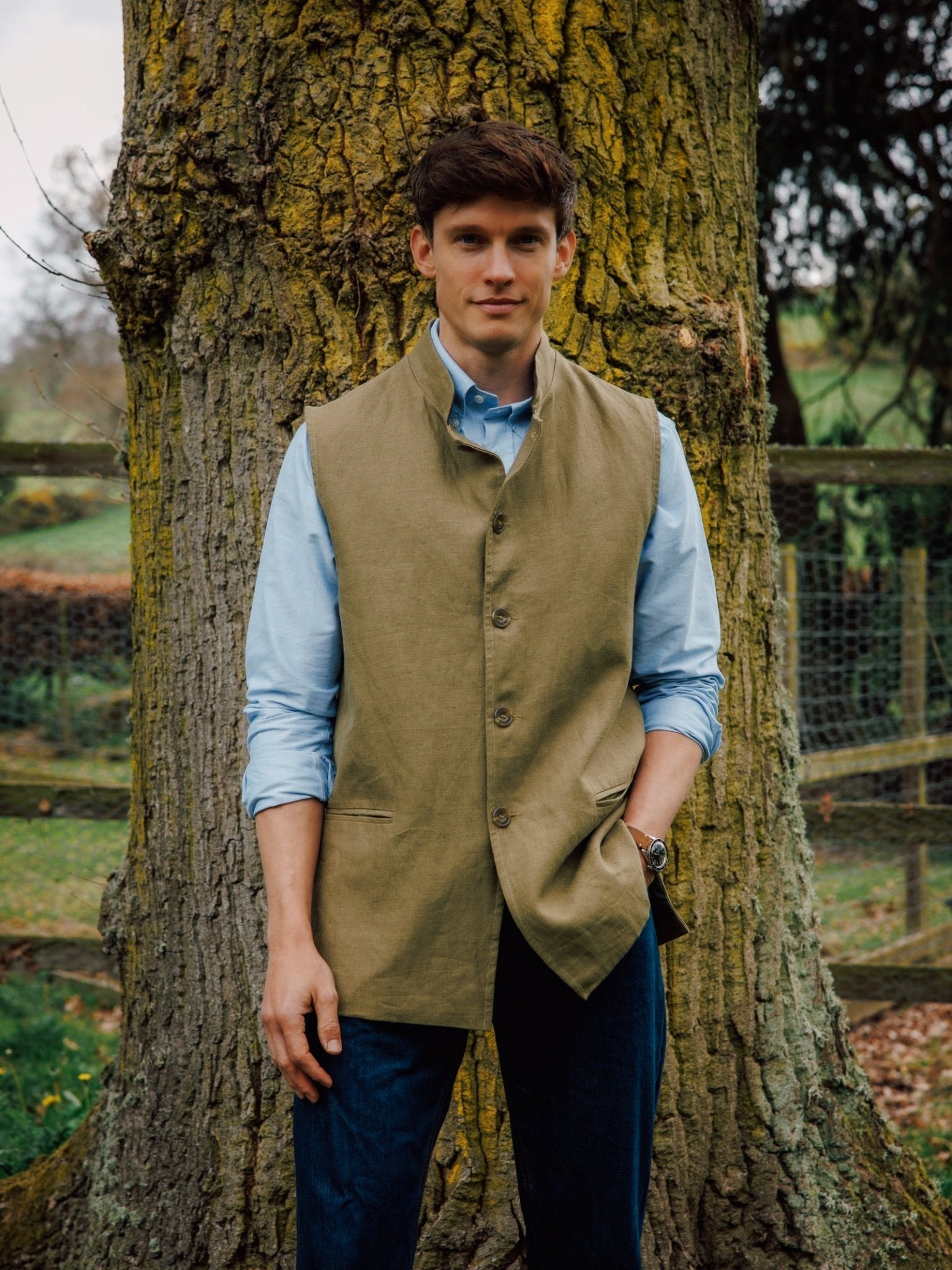 A man in a light blue shirt and a Campbell's of Beauly Linen Nehru Gilet stands outdoors by a large tree trunk, with greenery and a fence behind—an ideal refined summer layer.