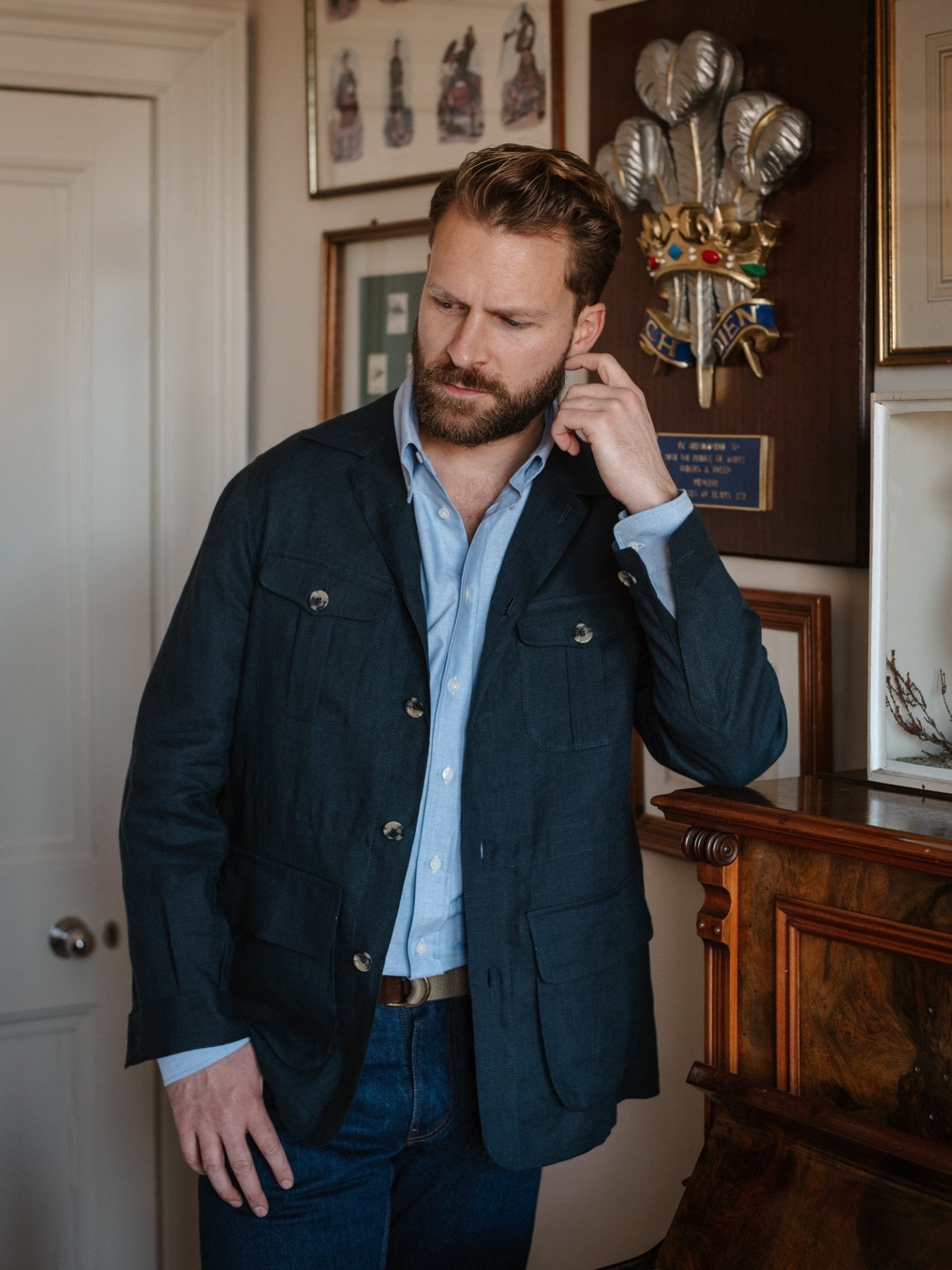 A bearded man wears the Campbell's of Beauly Linen Safari Jacket over a light blue shirt, leaning indoors by a wooden mantle among framed art—a stylish, refined layer for casual comfort.