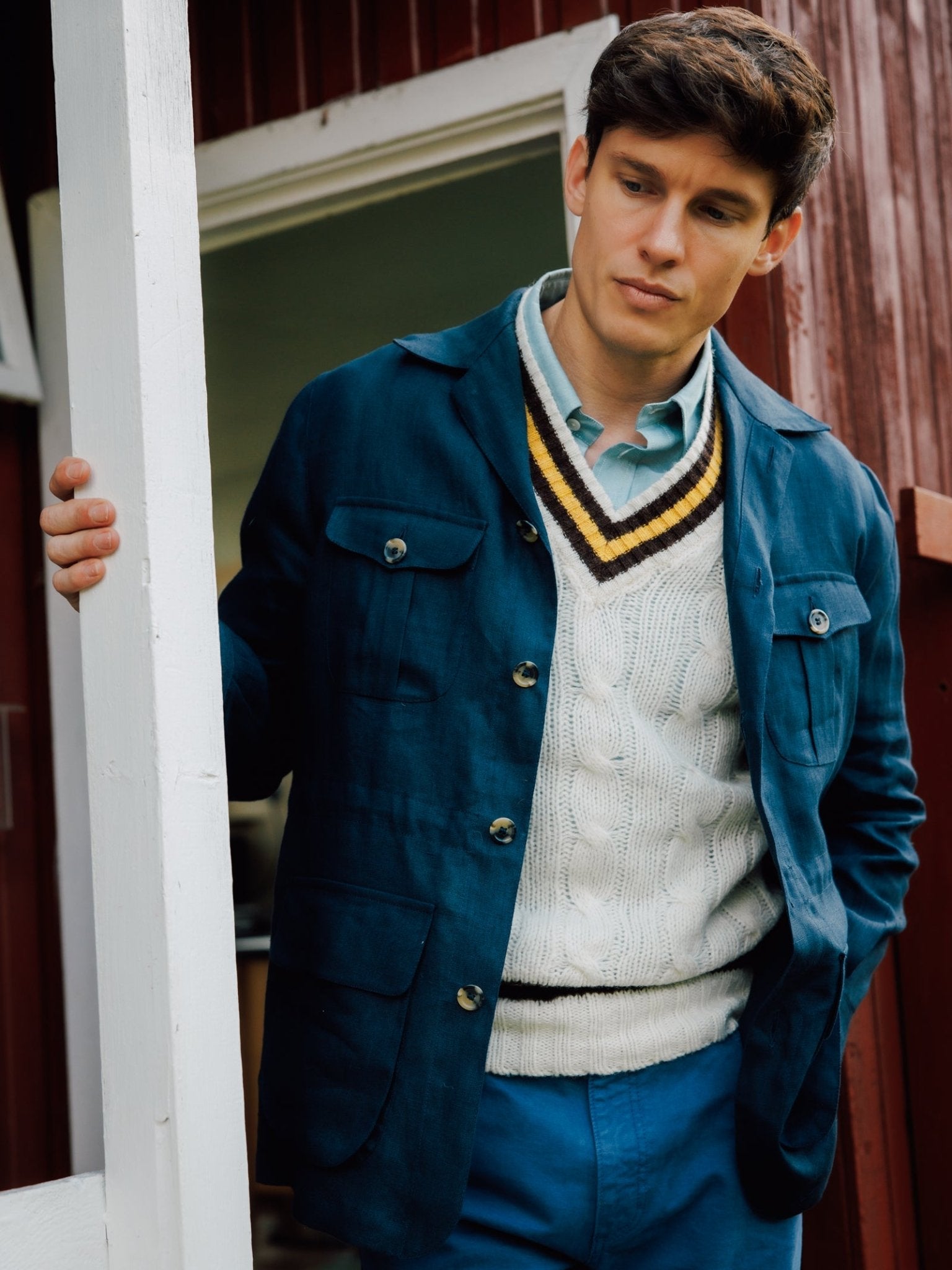A man in a blue Campbell's of Beauly Linen Safari Jacket over a white cable-knit sweater and blue pants stands in a doorway, looking down with one hand in his pocket—the breathable linen adds effortless style to his look.