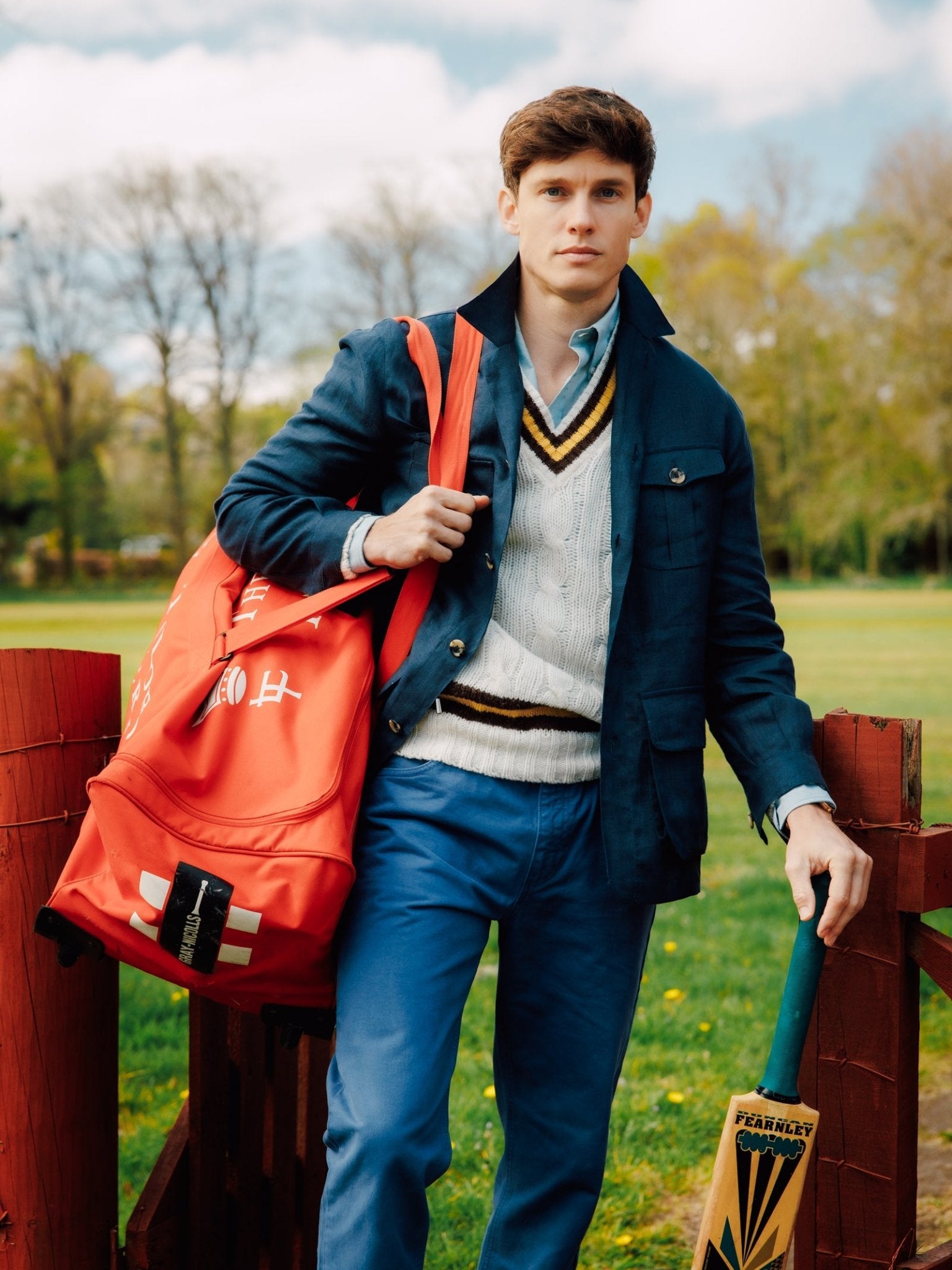 A young man in blue trousers, a sweater, and the Campbell's of Beauly Linen Safari Jacket stands outdoors with a red sports bag and cricket bat amid green trees—the perfect casual, breathable linen layer.