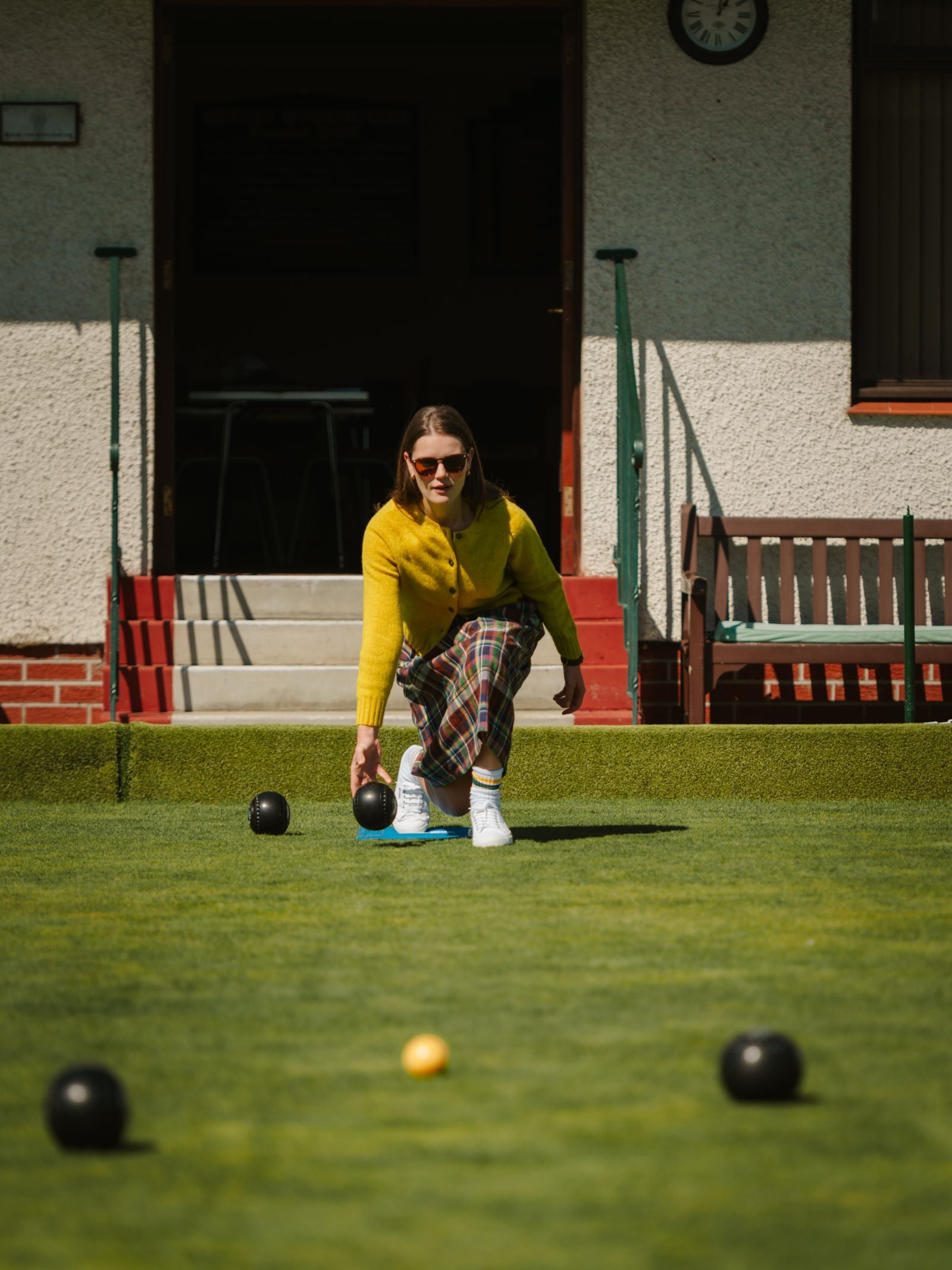 Wearing a Campbell's of Beauly Linen Skirt, a woman in a yellow sweater prepares to roll a black lawn bowl toward the yellow jack on a grassy lawn, with benches and a building in the background.