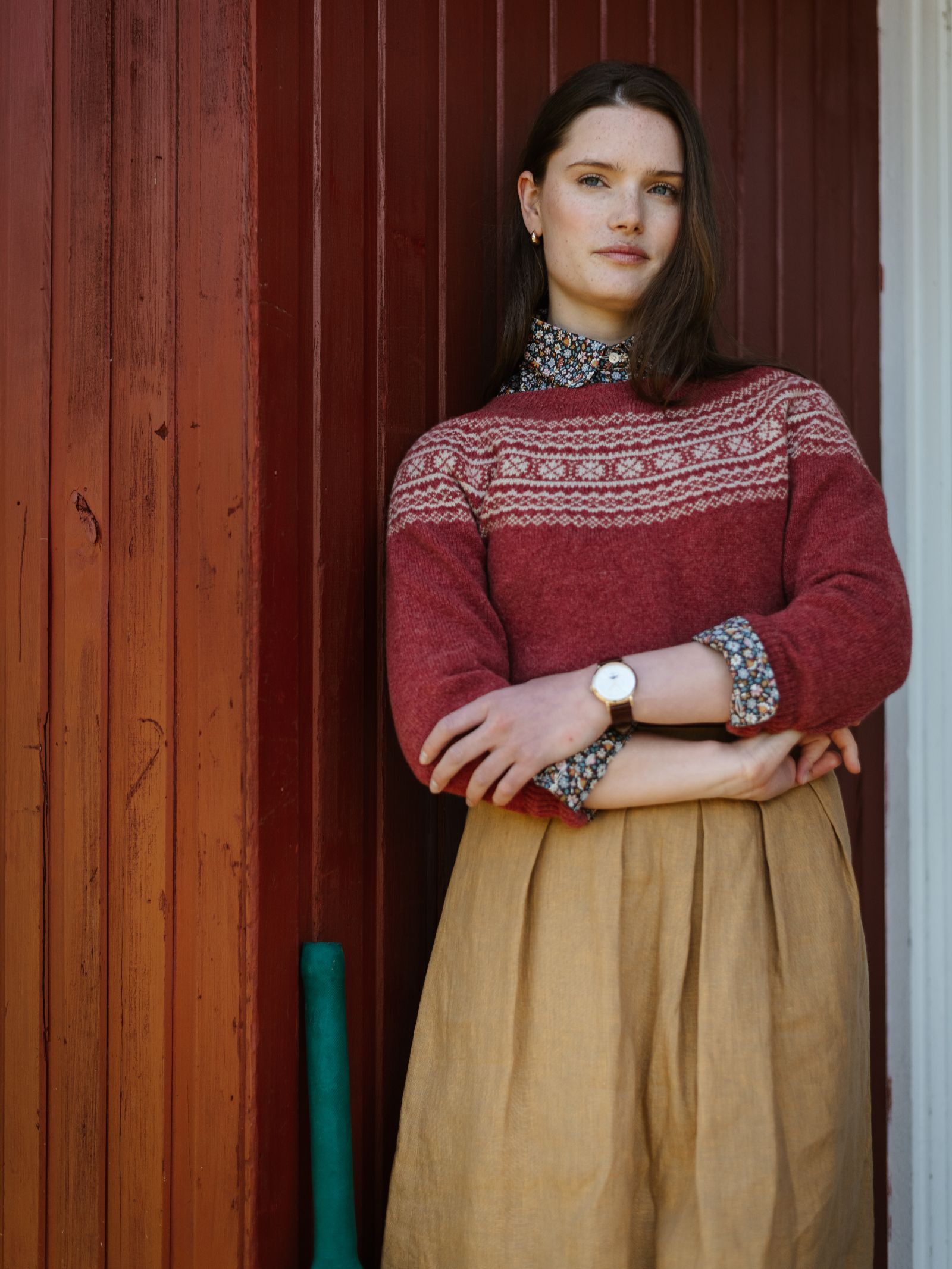 A woman with straight brown hair stands against a red wooden wall, wearing a patterned red sweater over a floral blouse and the Linen Skirt by Campbell's of Beauly. She crosses her arms and looks at the camera, with a green object beside her.