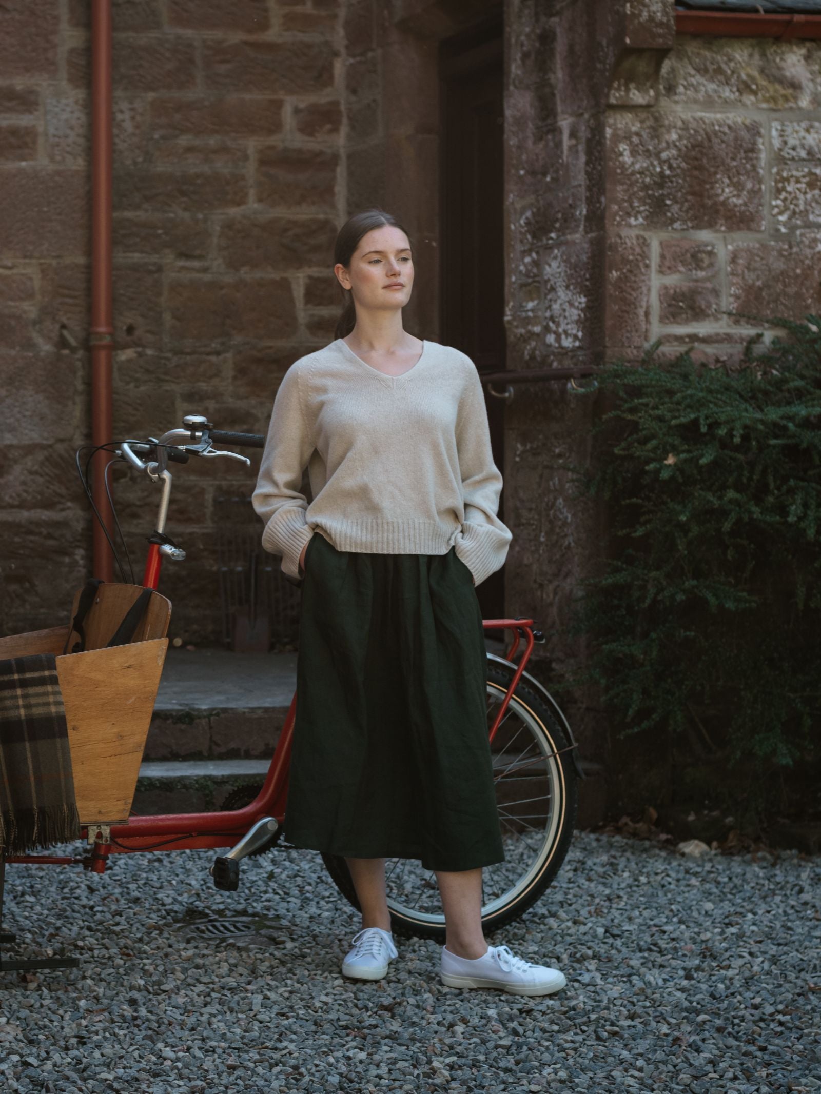 A woman stands on gravel beside a red cargo bike, wearing the Campbell's of Beauly Linen Skirt, a beige sweater, and white sneakers. Her hands are in her pockets; behind her is a brick wall with greenery.