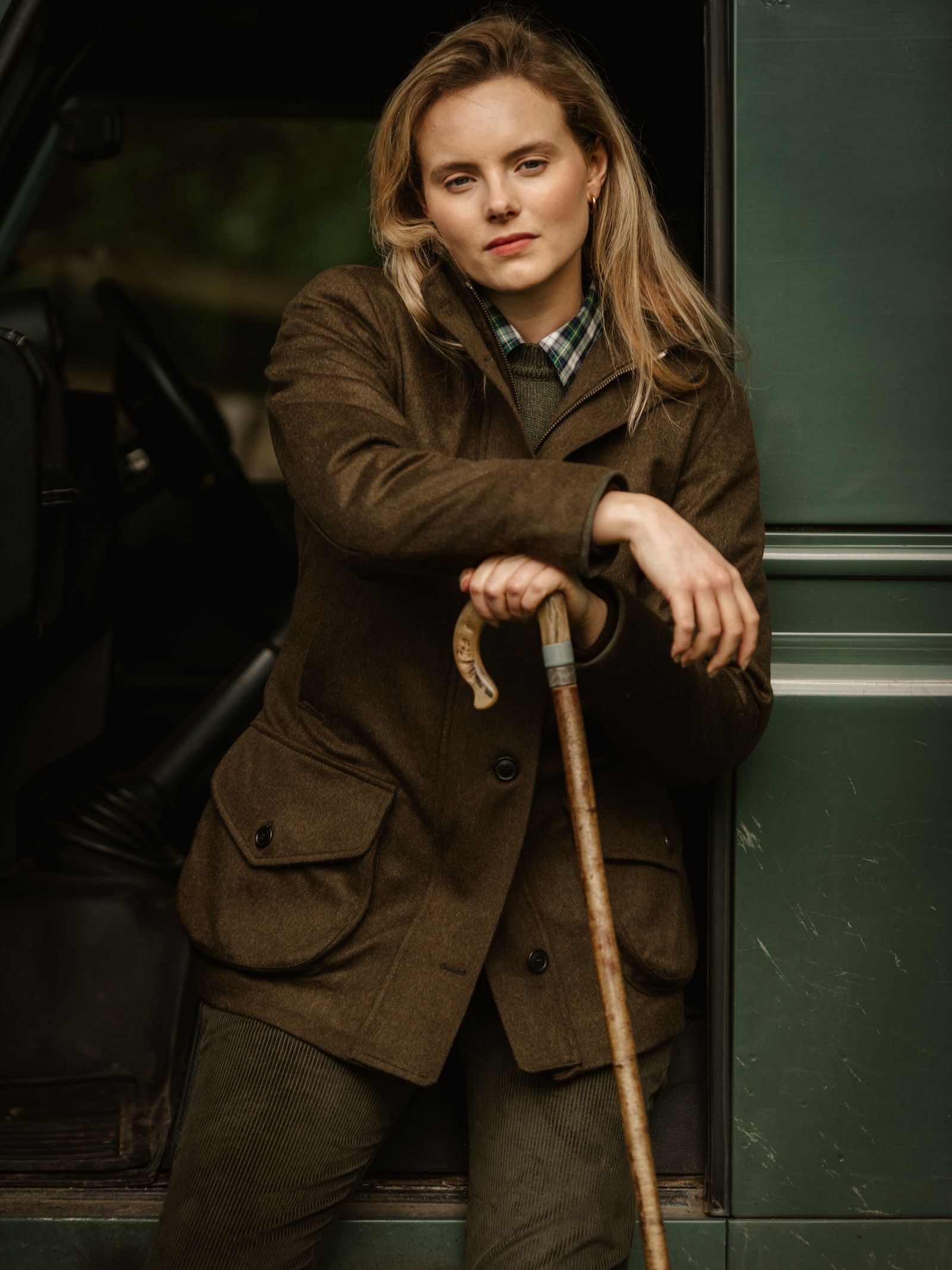 A woman wears the Campbell's of Beauly Loden Green Field Coat, crafted in the UK from classic Austrian loden fabric, over a plaid shirt as she leans on a walking stick beside an open green vehicle, gazing confidently at the camera.