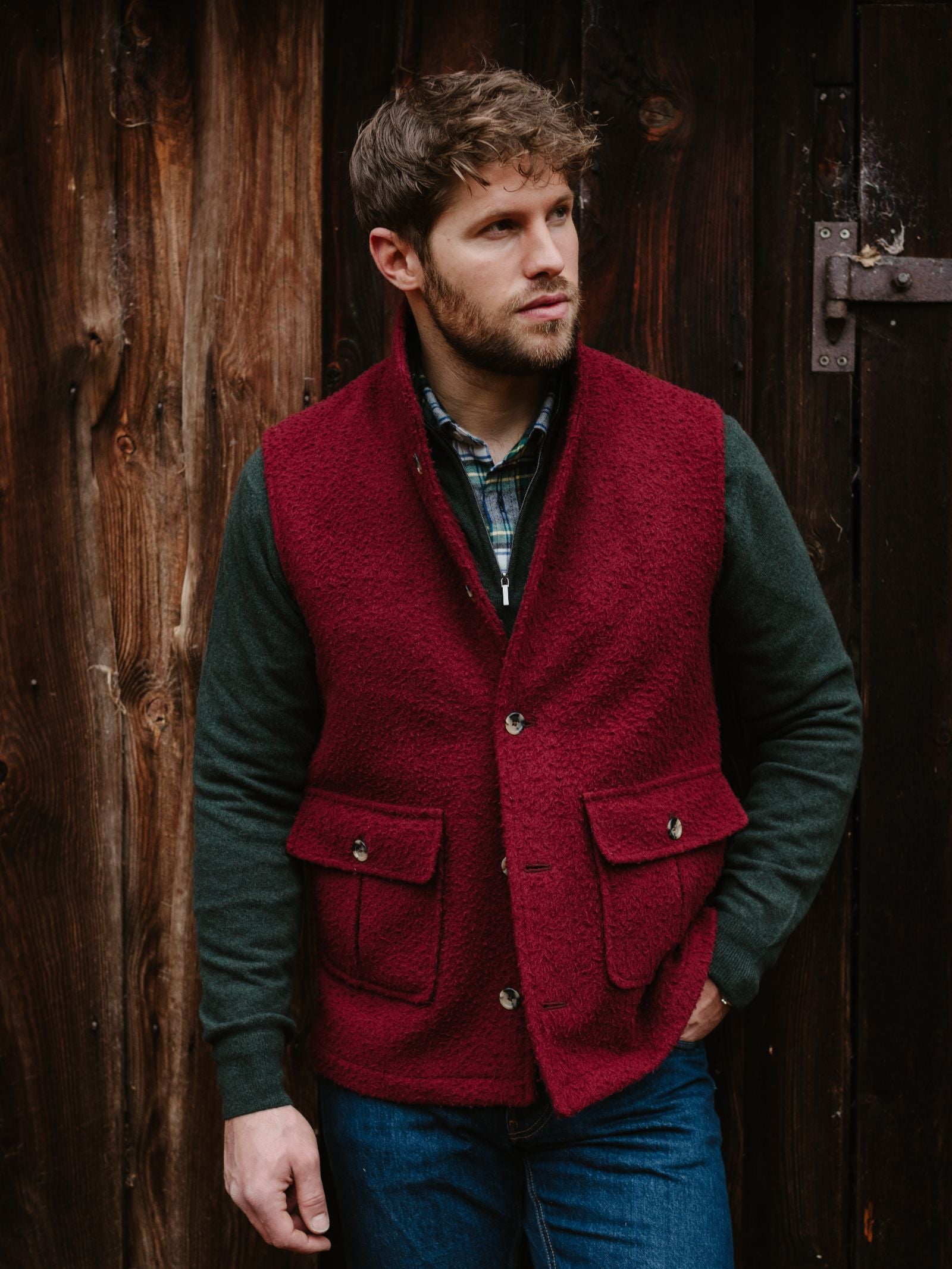 A man with wavy brown hair and a beard wears a dark green shirt and a red Campbell's of Beauly Casentino Fleece Gilet with pockets, standing before a rustic wooden wall with a visible metal hinge.