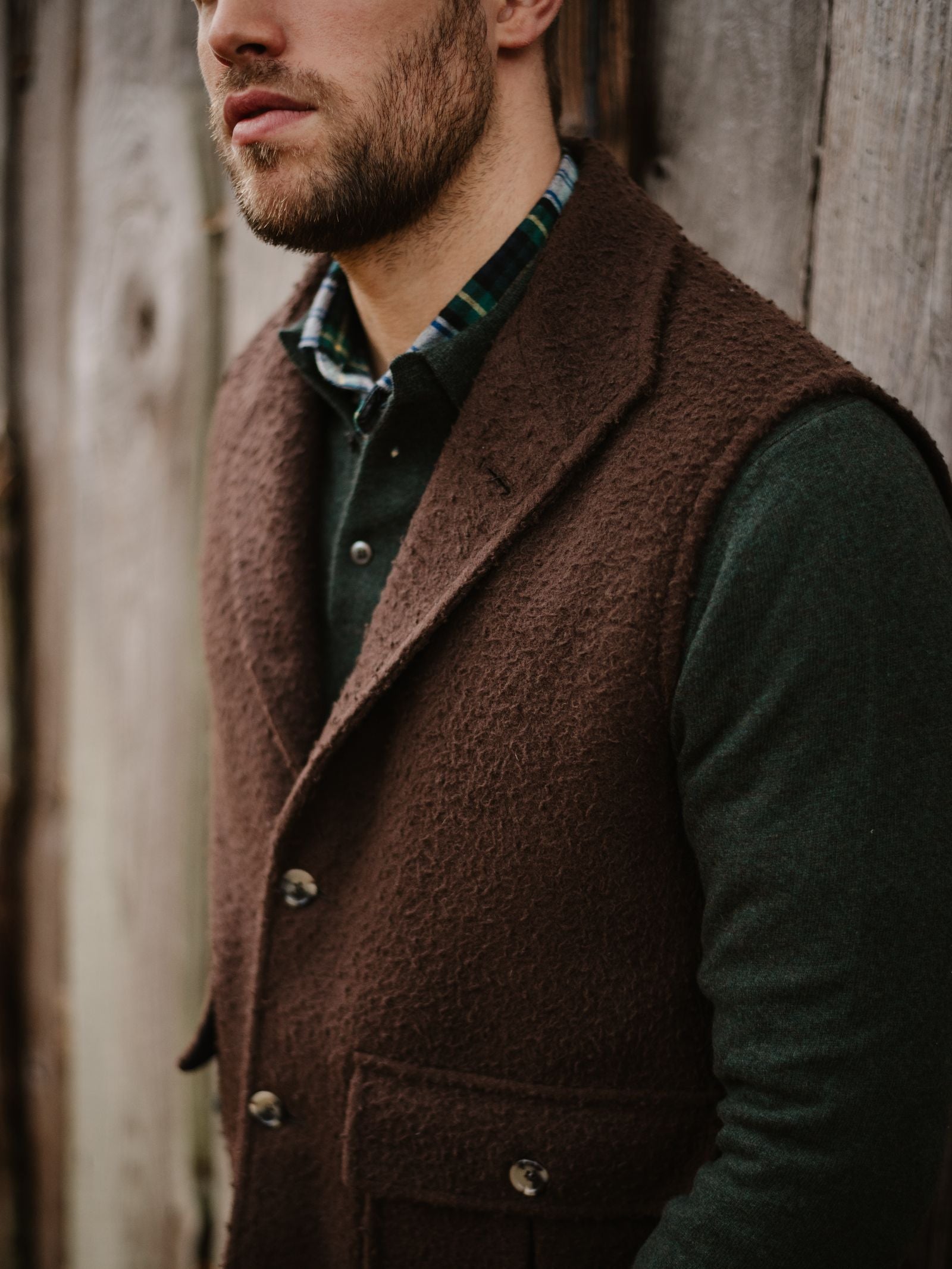 A man wearing a dark green shirt layered with a brown Campbell's of Beauly Casentino Fleece Gilet stands before a weathered wooden wall, with only the lower half of his face visible.