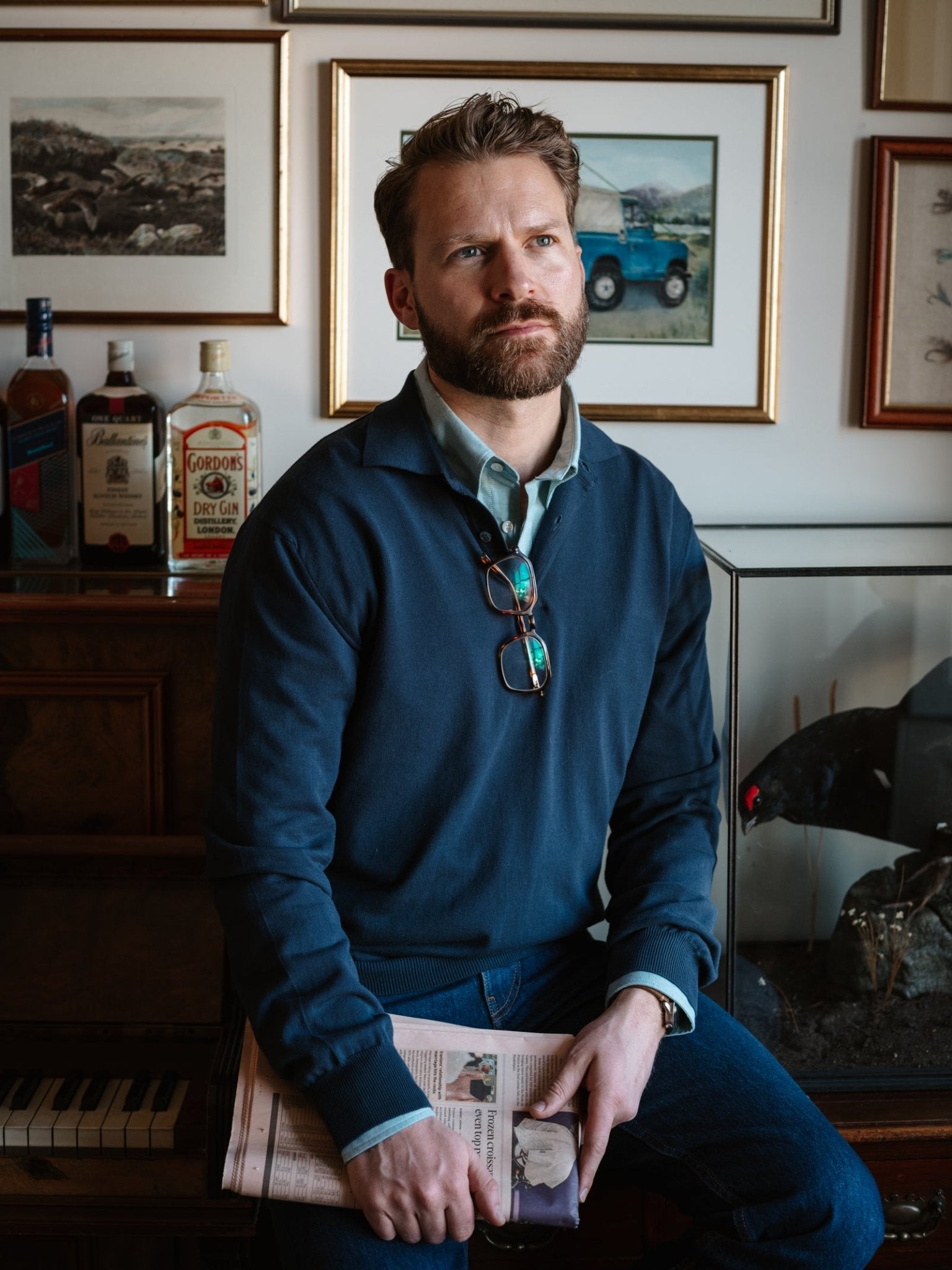 A bearded man in a blue Campbell's of Beauly Cotton Rugby sits on a piano bench holding a newspaper, glasses around his neck. He gazes thoughtfully into the distance, with bottles, framed pictures, and a glass tank behind him.