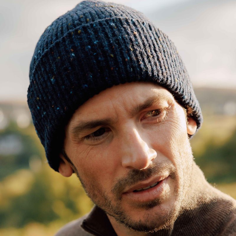 A man with light stubble wears the Campbells of Beauly Donegal Lambswool Hat, a blue speckled knit beanie, paired with a brown jacket as he looks to the side outdoors against a blurred, sunlit background.