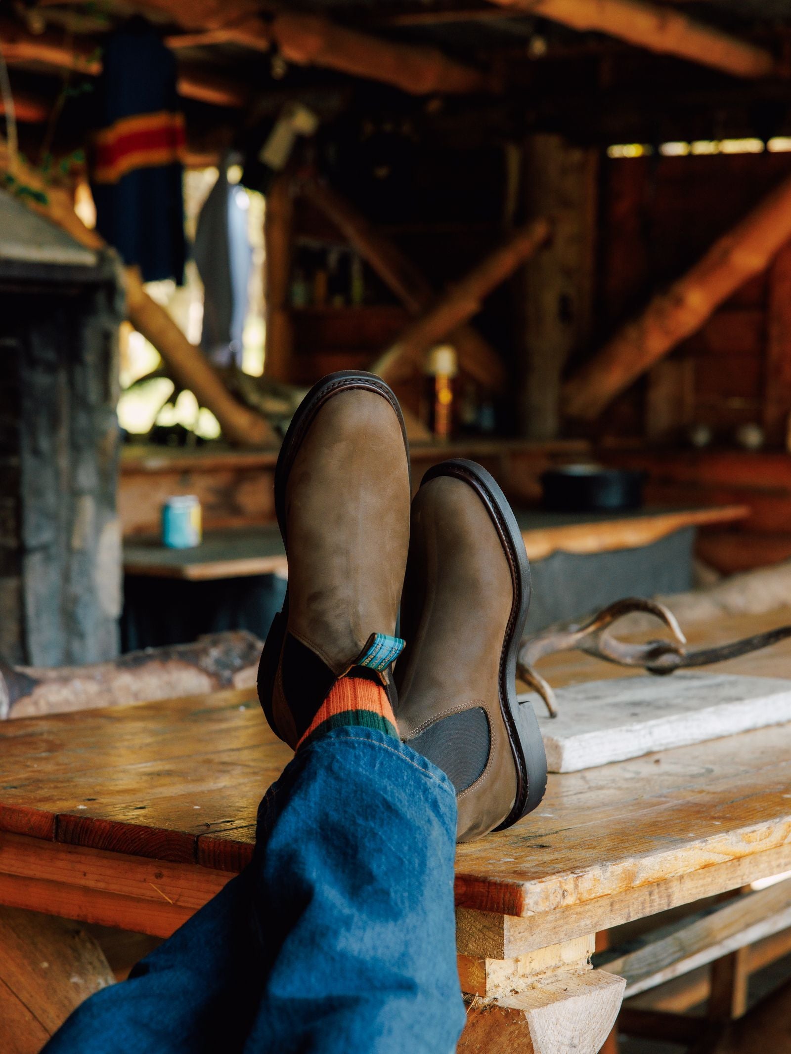 Relaxing in a cozy wooden cabin, a person lounges with feet up on a rustic table, showcasing Men's R.M.Williams Highlander Boots by R.M.Williams x Campbell's of Beauly, paired with orange socks and blue jeans amid wooden beams and hanging clothes.