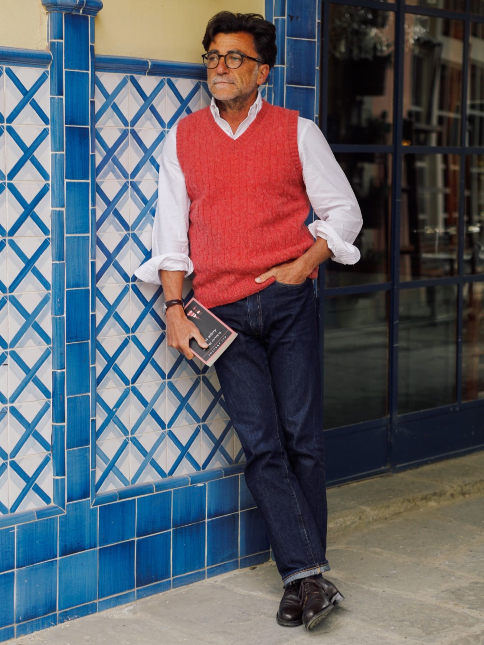 A man in glasses and a white shirt wears the Campbell's of Beauly Shetland Cable Slipover in red. He stands casually against a blue-tiled wall holding a book, dressed in dark jeans and black shoes with one leg crossed over the other.