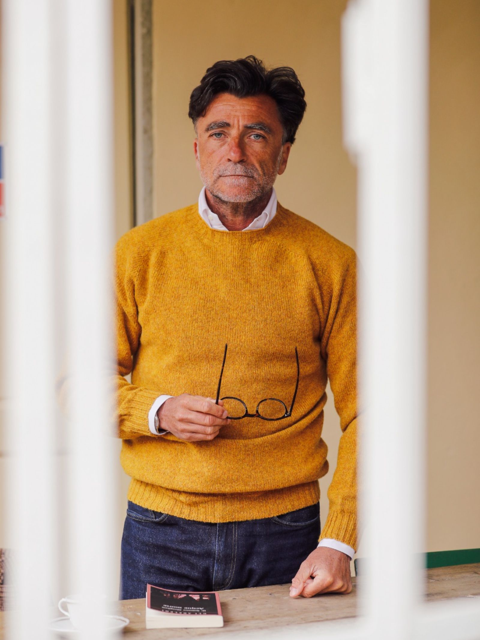 A man with dark hair and a beard, wearing the Shetland Jumper in mustard yellow by Campbell's of Beauly and jeans, stands indoors holding eyeglasses. A book and cup sit on the table as he looks at the camera.