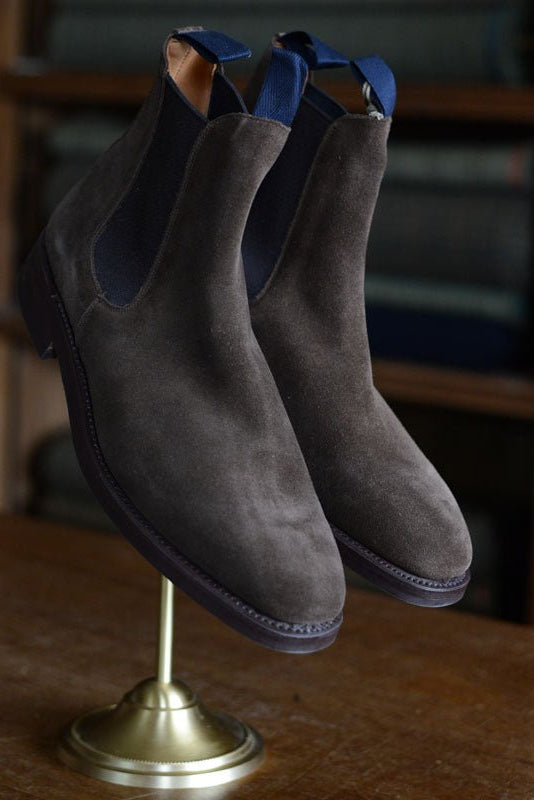 Campbell's of Beauly Auld Stock Suede Chelsea Boots in dark brown feature blue pull tabs and a rugged commando sole, shown on a brass stand with blurred fabric rolls behind—a classic British design.