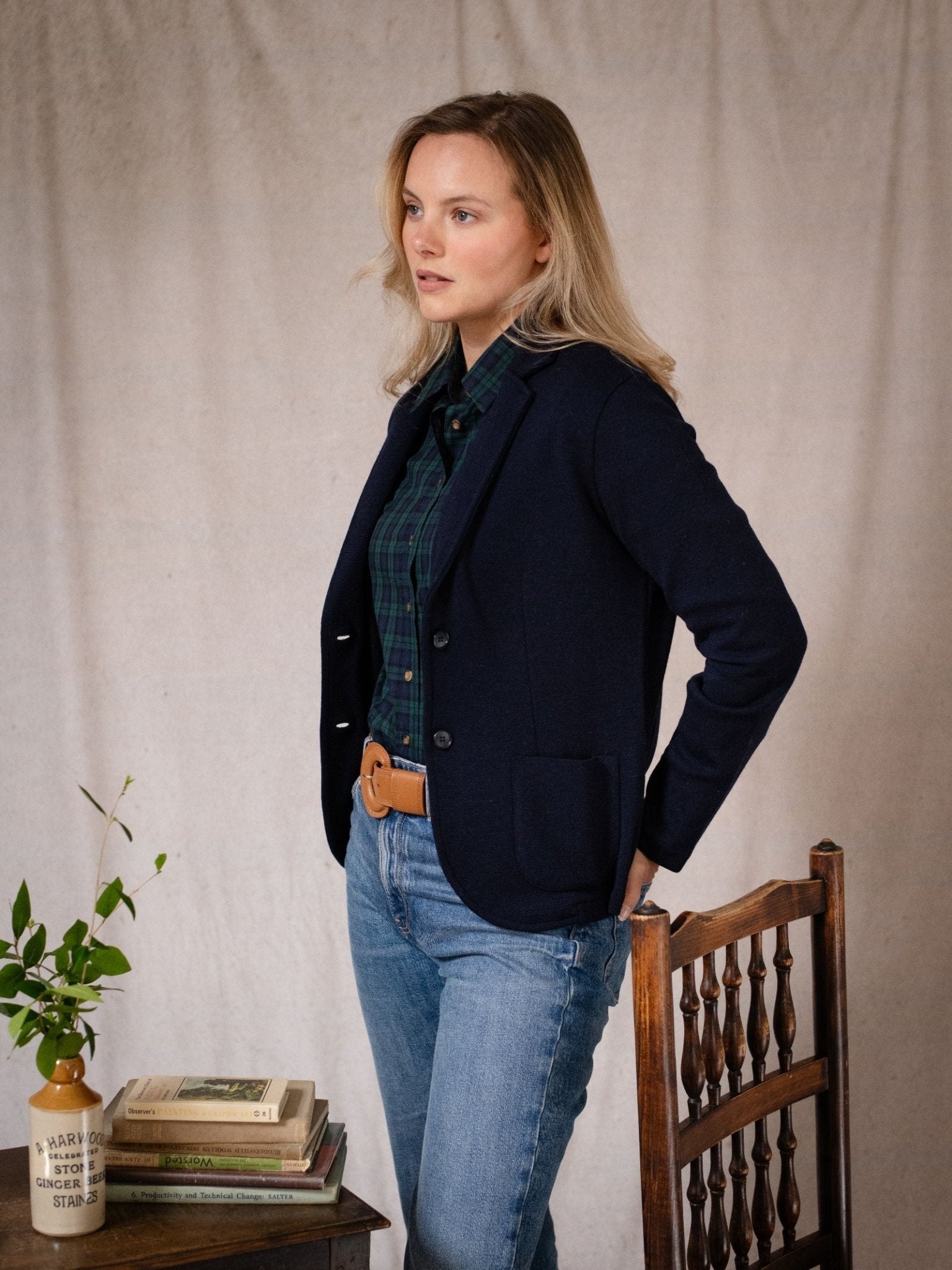 A blonde woman stands indoors in a navy Campbell's of Beauly Merino Blazer over a green plaid shirt and jeans, beside a wooden chair and table with books and a vase of green branches, with a neutral fabric backdrop behind her.