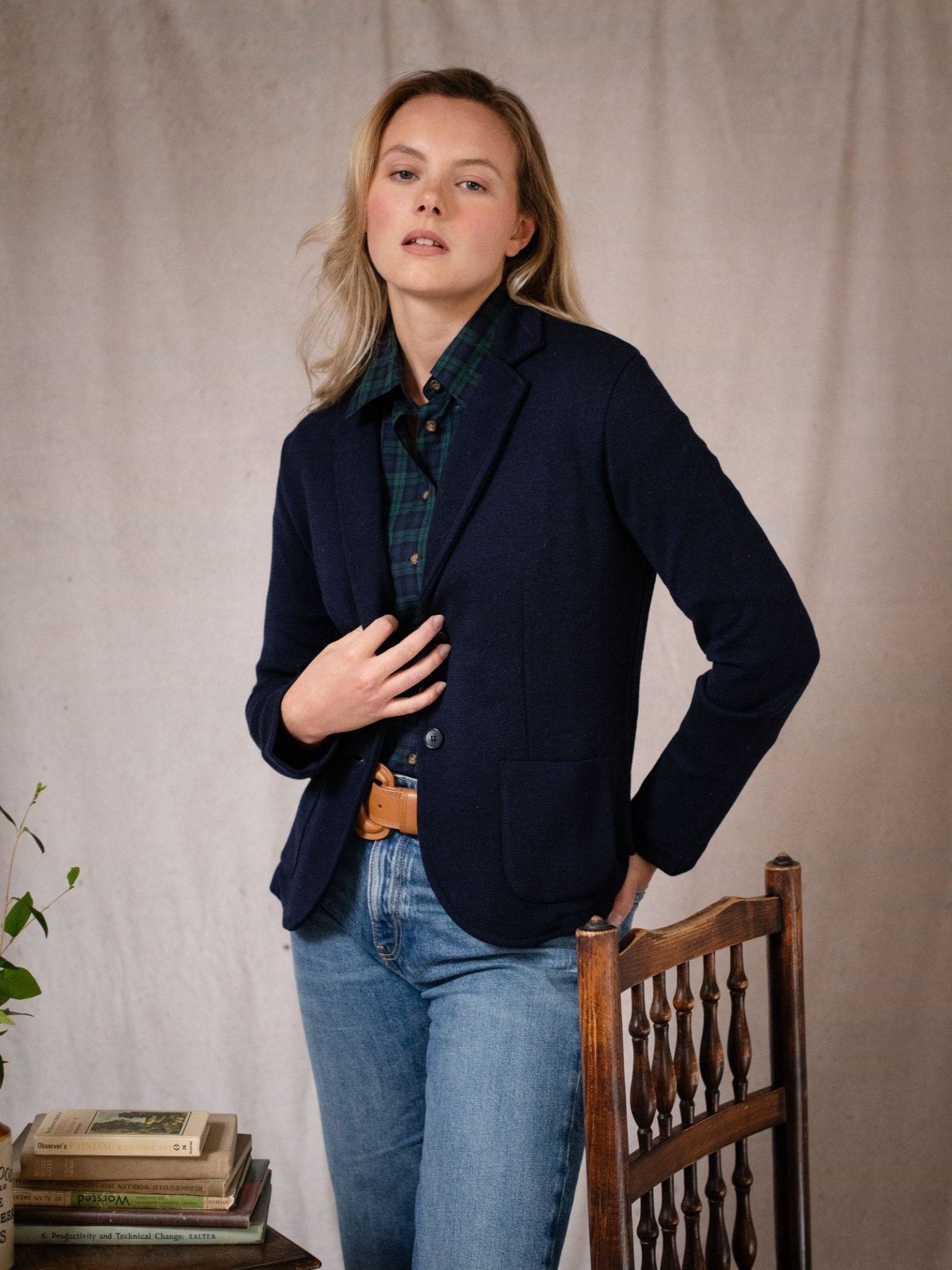 A blonde woman wears the Campbell’s of Beauly Merino Blazer in navy over a green plaid shirt, standing by a wooden chair with her hand on her chest. The blazer is made from fine merino wool for elegance and comfort.