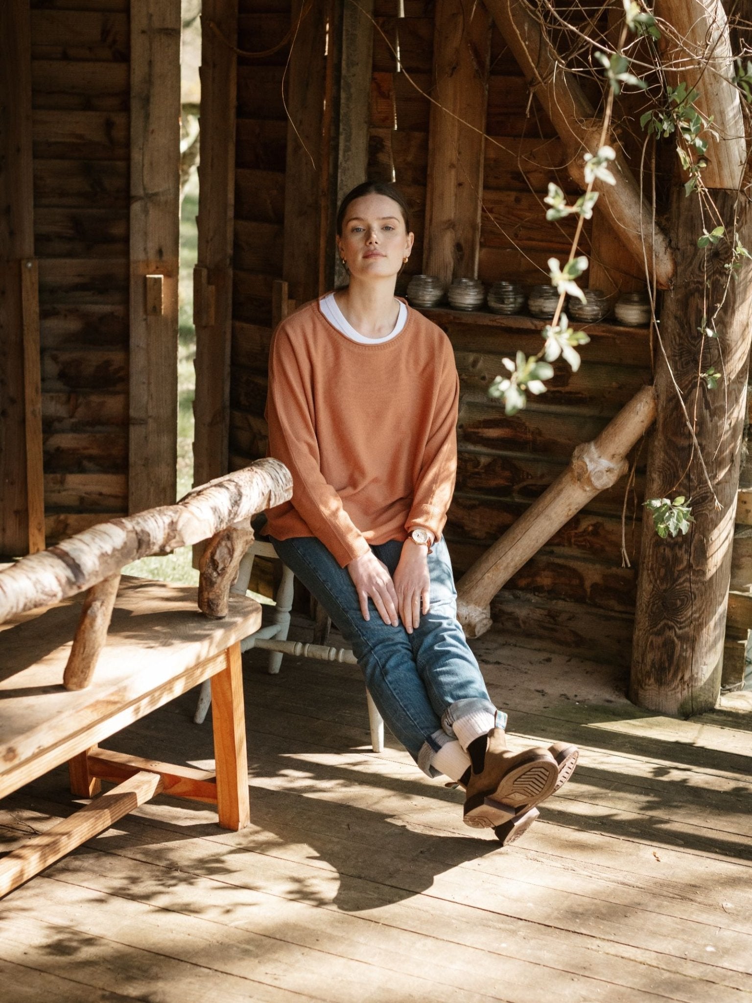 A person in a Campbell’s of Beauly Merino Cashmere Boat Neck Jumper, jeans, and an orthopedic leg brace sits on a white bench inside a rustic wooden cabin, sunlight streaming in and greenery visible outside.