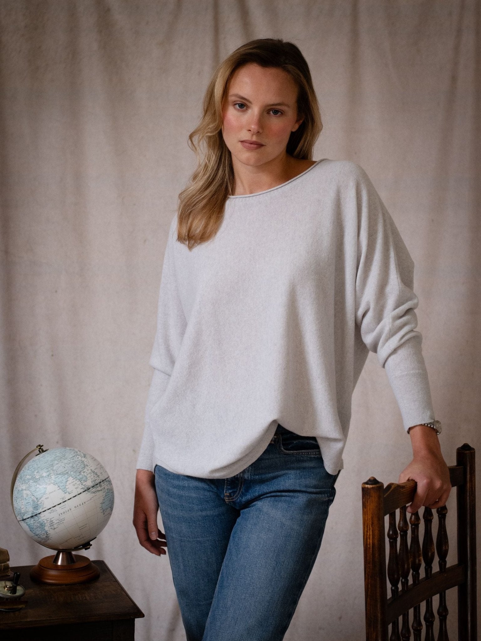 A woman with long blonde hair wears the Campbell's of Beauly Merino Cashmere Boat Neck Jumper and jeans, standing indoors by a wooden chair. A small globe rests on a table beside her against a plain beige fabric background.