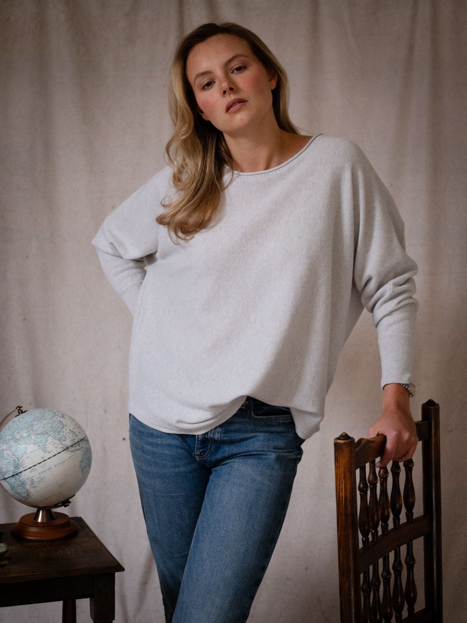 A woman with long blonde hair wears the Campbell’s of Beauly Merino Cashmere Boat Neck Jumper and blue jeans, leaning against a wooden chair beside a table with a globe, set against a neutral fabric backdrop.