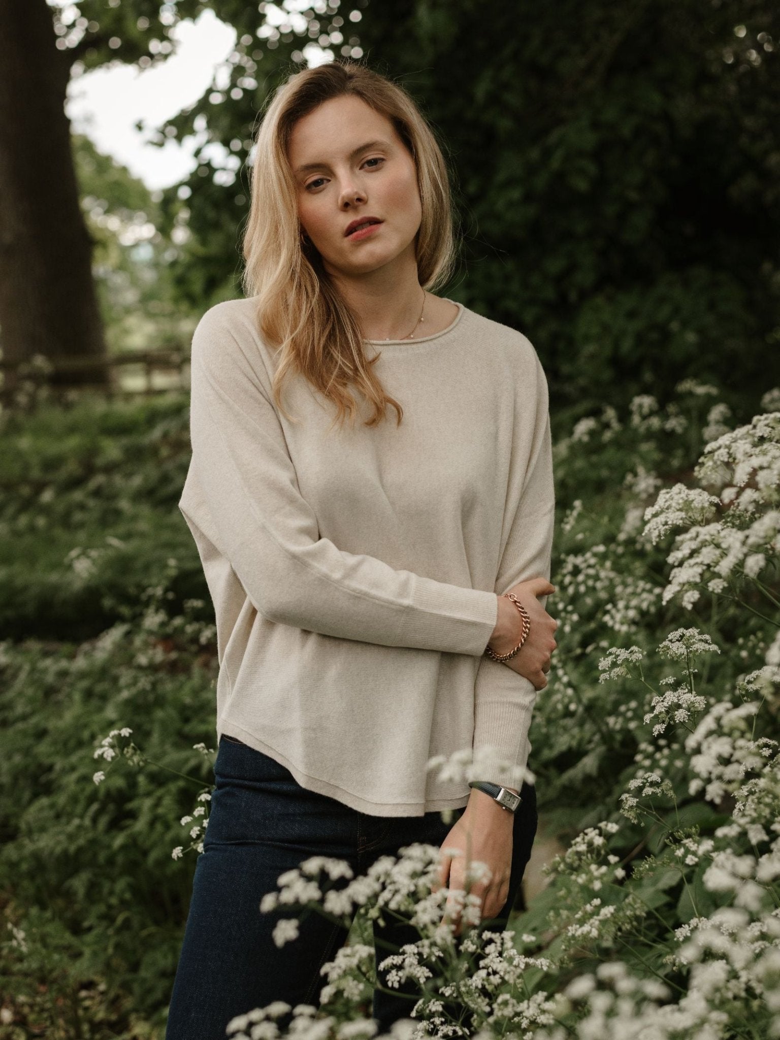 A woman with long blonde hair stands among white wildflowers and greenery, wearing the Campbell's of Beauly Merino Cashmere Boat Neck Jumper in light beige and dark jeans. She appears relaxed and thoughtful.