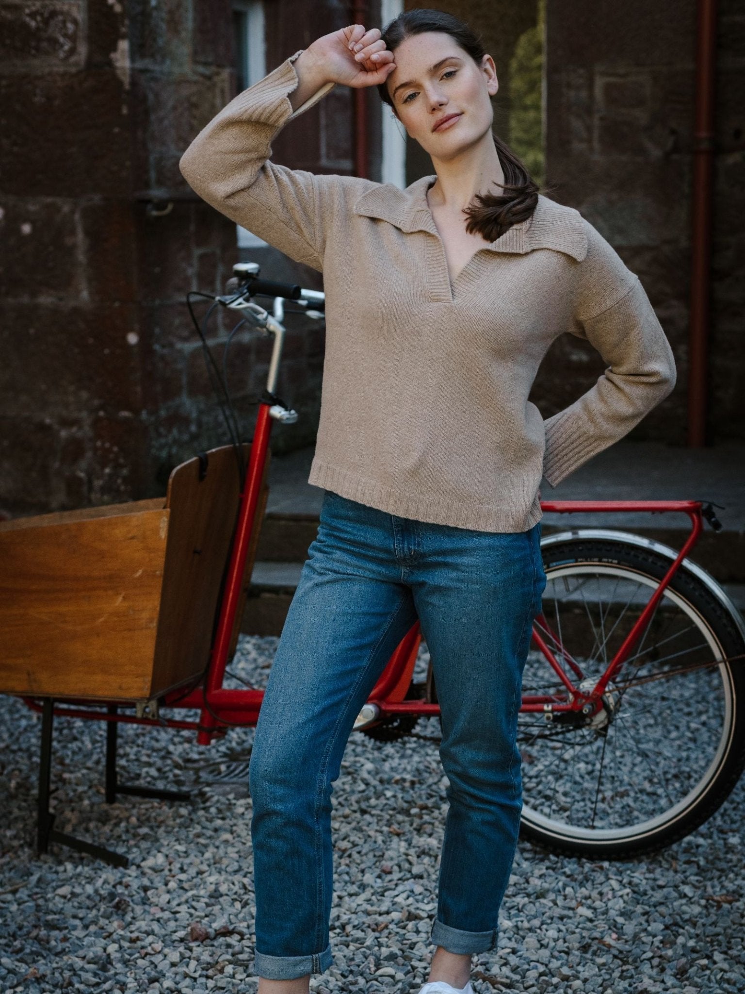 A woman wears the Campbell’s of Beauly Merino Cashmere Shirt Neck Jumper in beige with blue jeans, standing before a red bicycle with a wooden cargo box, one arm raised, against a stone wall and gravel ground.