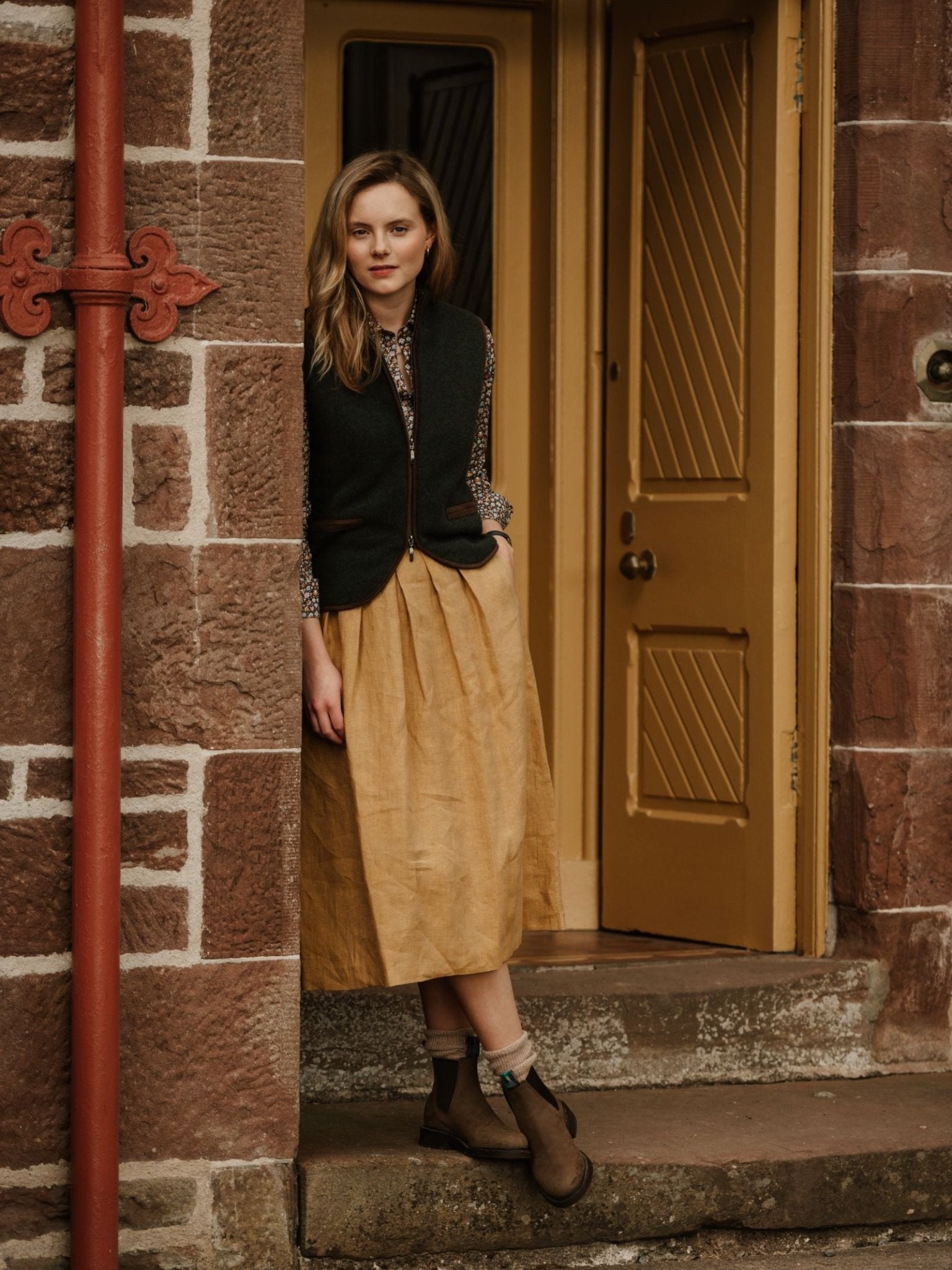 A woman with light brown hair stands in a doorway styled for the countryside, wearing Campbell's of Beauly Merino Cashmere Gilet over a patterned shirt, tan midi skirt, and brown ankle boots. Yellow door and reddish stone walls complete the look.