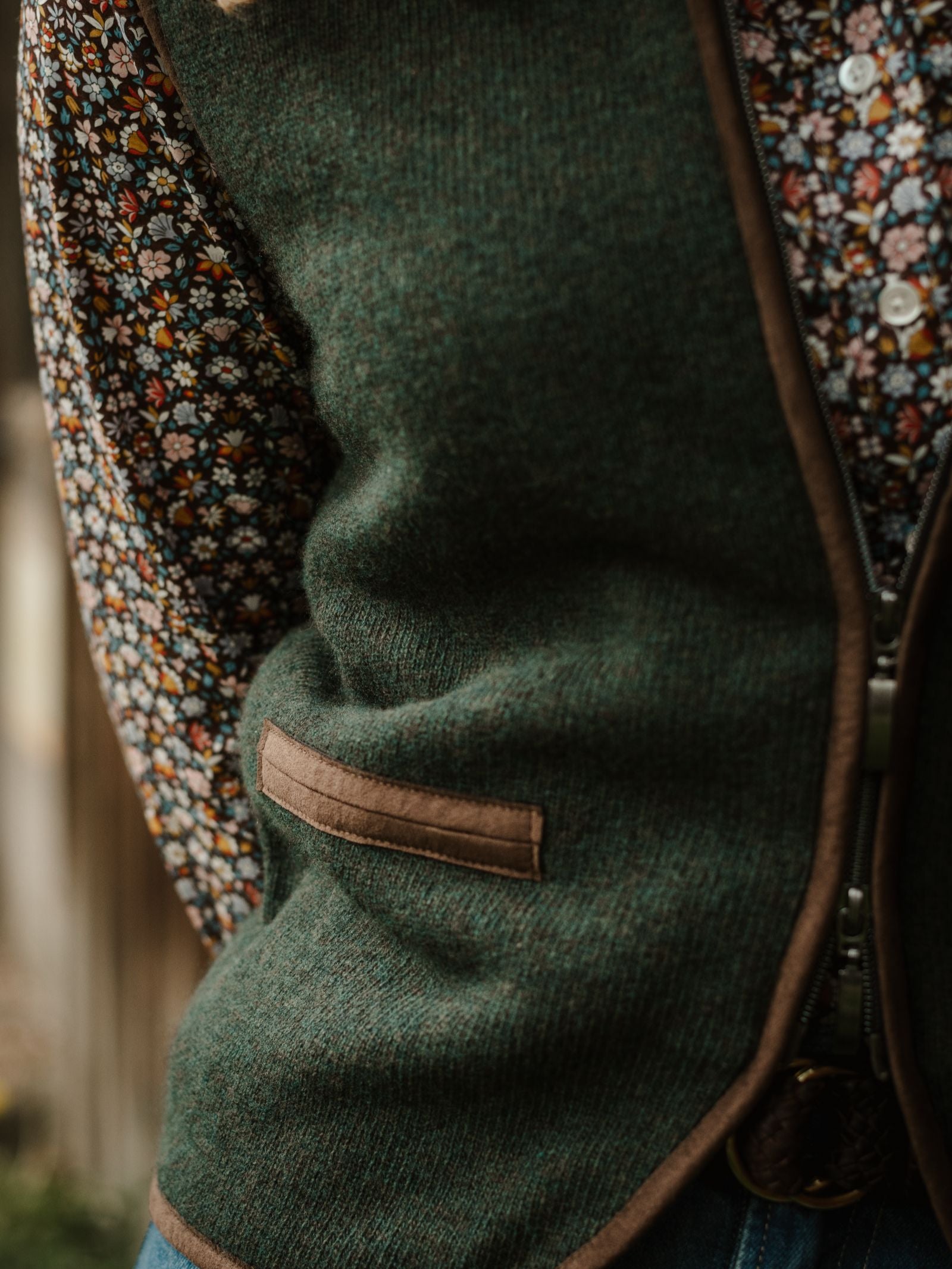 Close-up of a person wearing the Campbell's of Beauly Merino Cashmere Gilet, featuring brown trim and a pocket, layered over a vibrant floral shirt. The gilet's zipper is partly visible against a softly blurred countryside background.