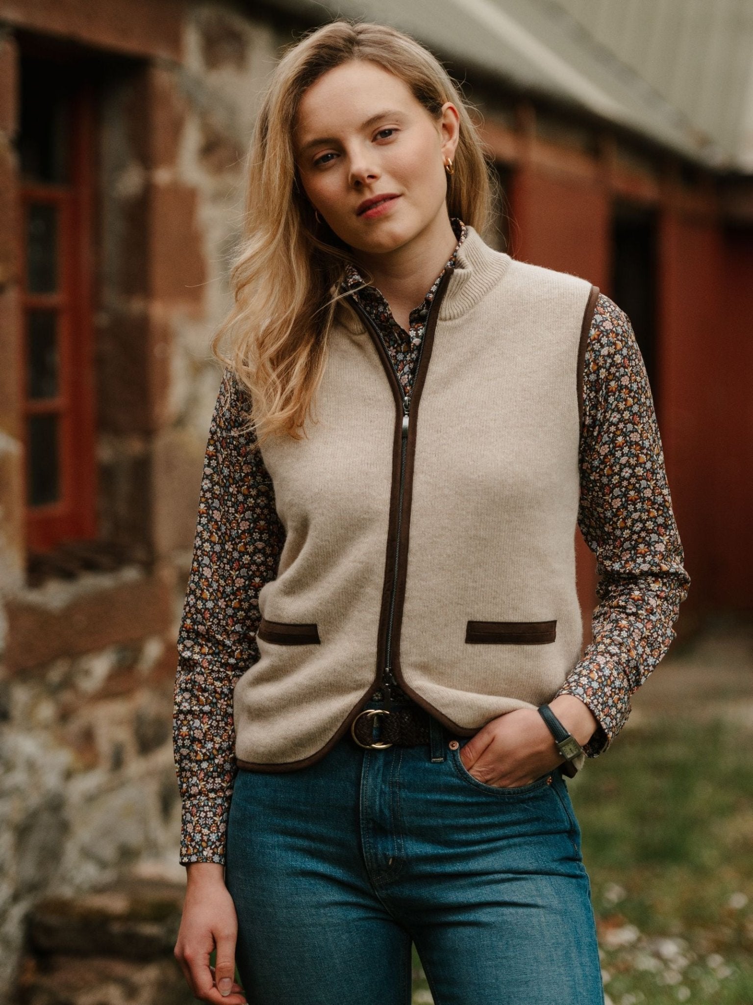 A young woman with blond hair, dressed in a Campbell's of Beauly Merino Cashmere Gilet over a floral shirt and blue jeans, stands confidently by a rustic stone building, embodying countryside style.