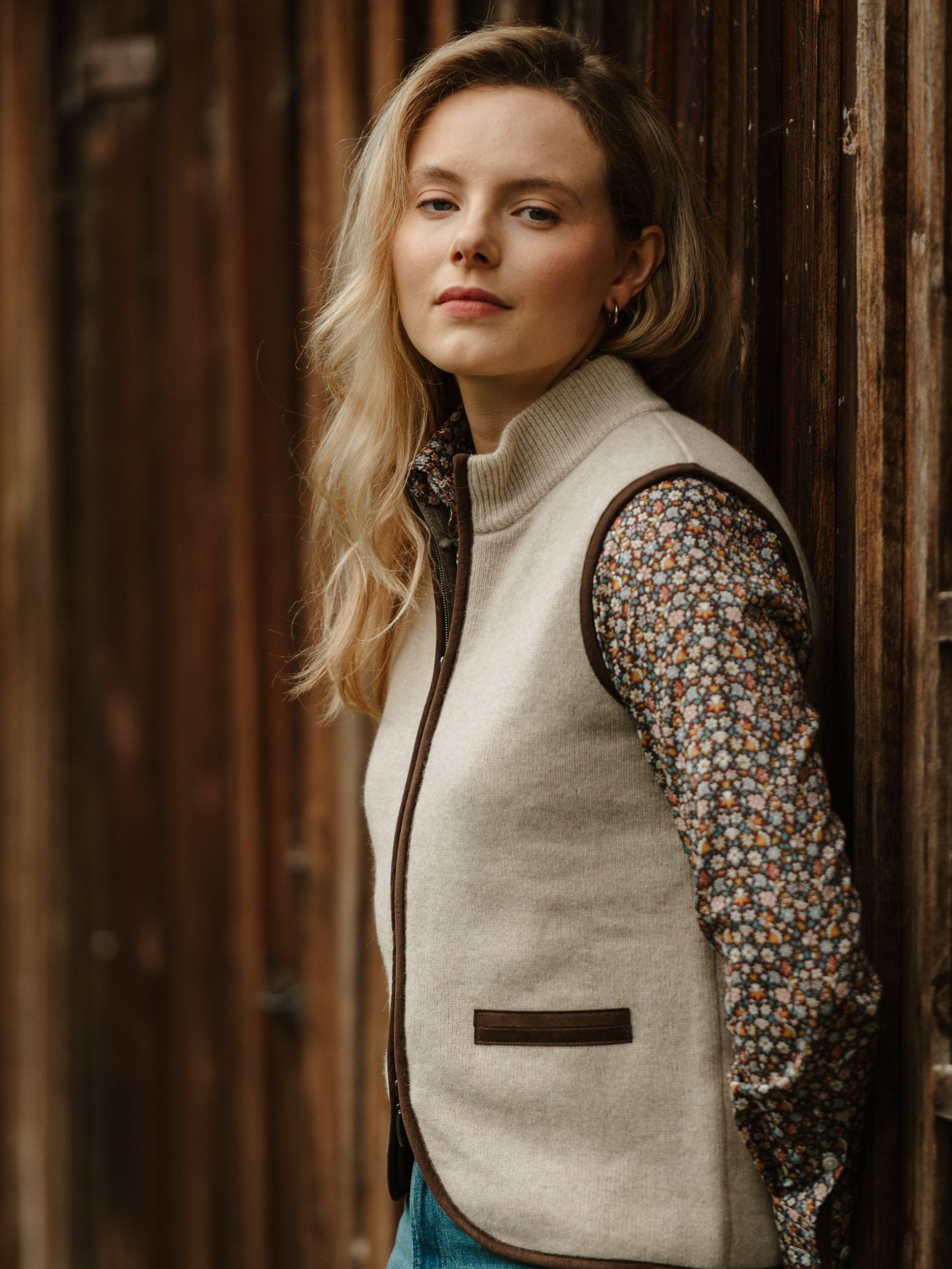 A woman with long blonde hair wears the Campbell's of Beauly Merino Cashmere Gilet over a floral shirt and leans against a wooden wall, looking calmly at the camera.