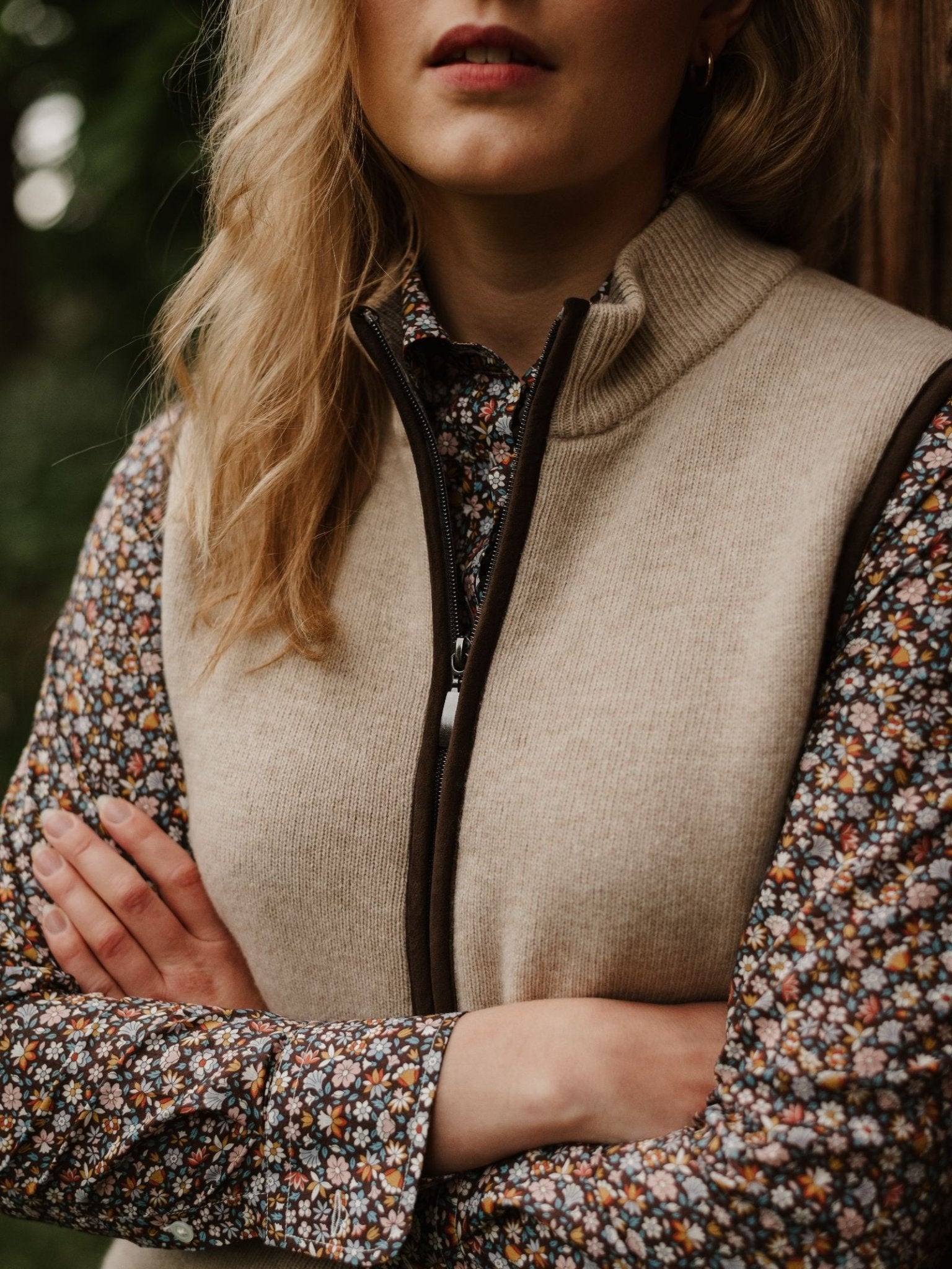 A woman with blonde hair, arms crossed, wears the Campbell's of Beauly Merino Cashmere Gilet over a floral long-sleeve shirt. She poses outdoors by a wooden surface with her face partially visible.