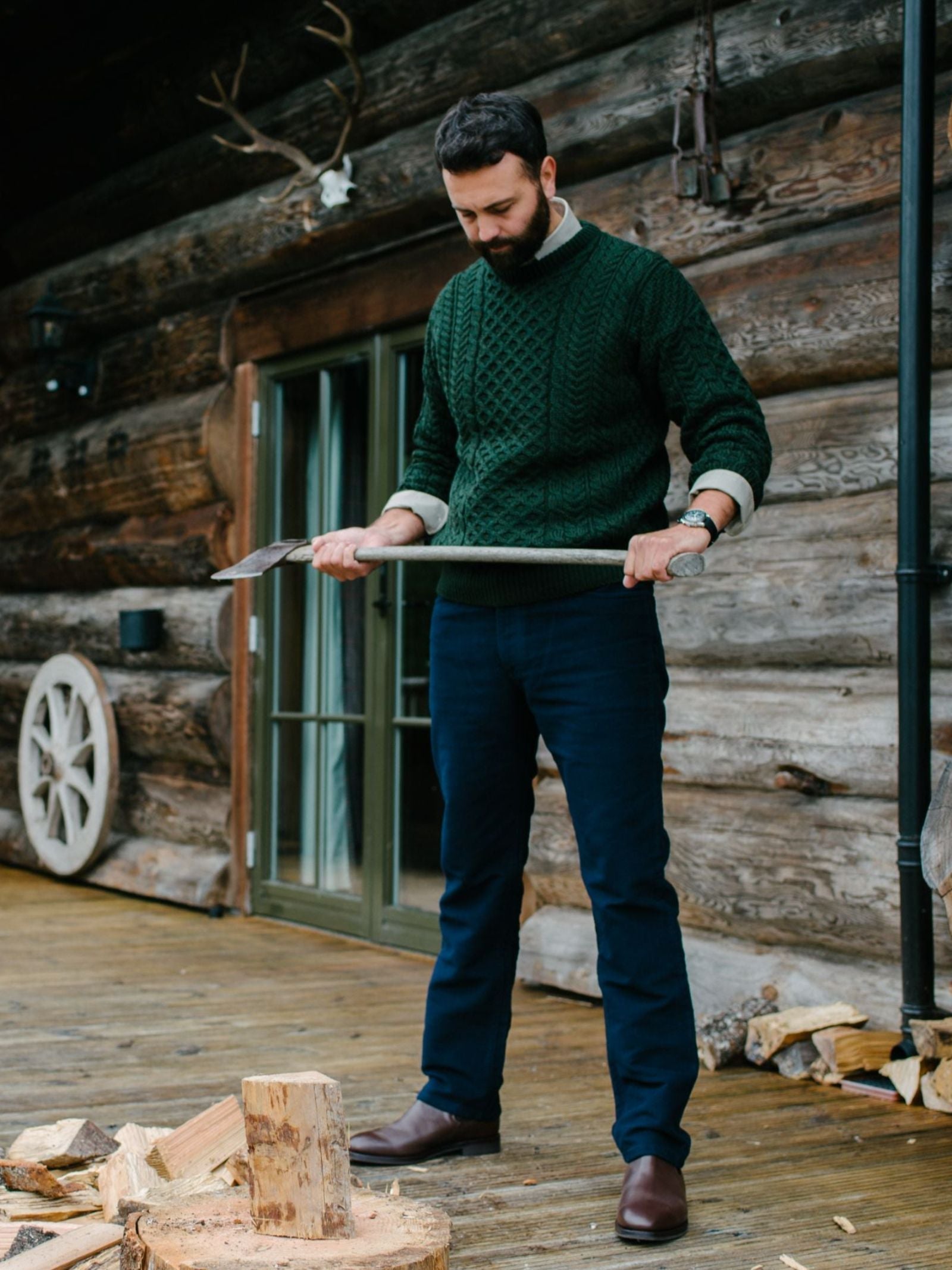 A bearded man stands on a wooden porch in front of a log cabin, holding an axe and preparing to chop firewood. He wears a green sweater, brown boots, and Campbell's of Beauly Moleskin Jeans. Logs are scattered nearby.